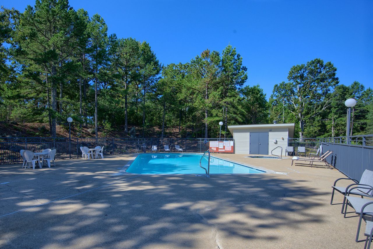 Swimming pool surrounded by trees, with chairs and a small building under a bright blue sky.