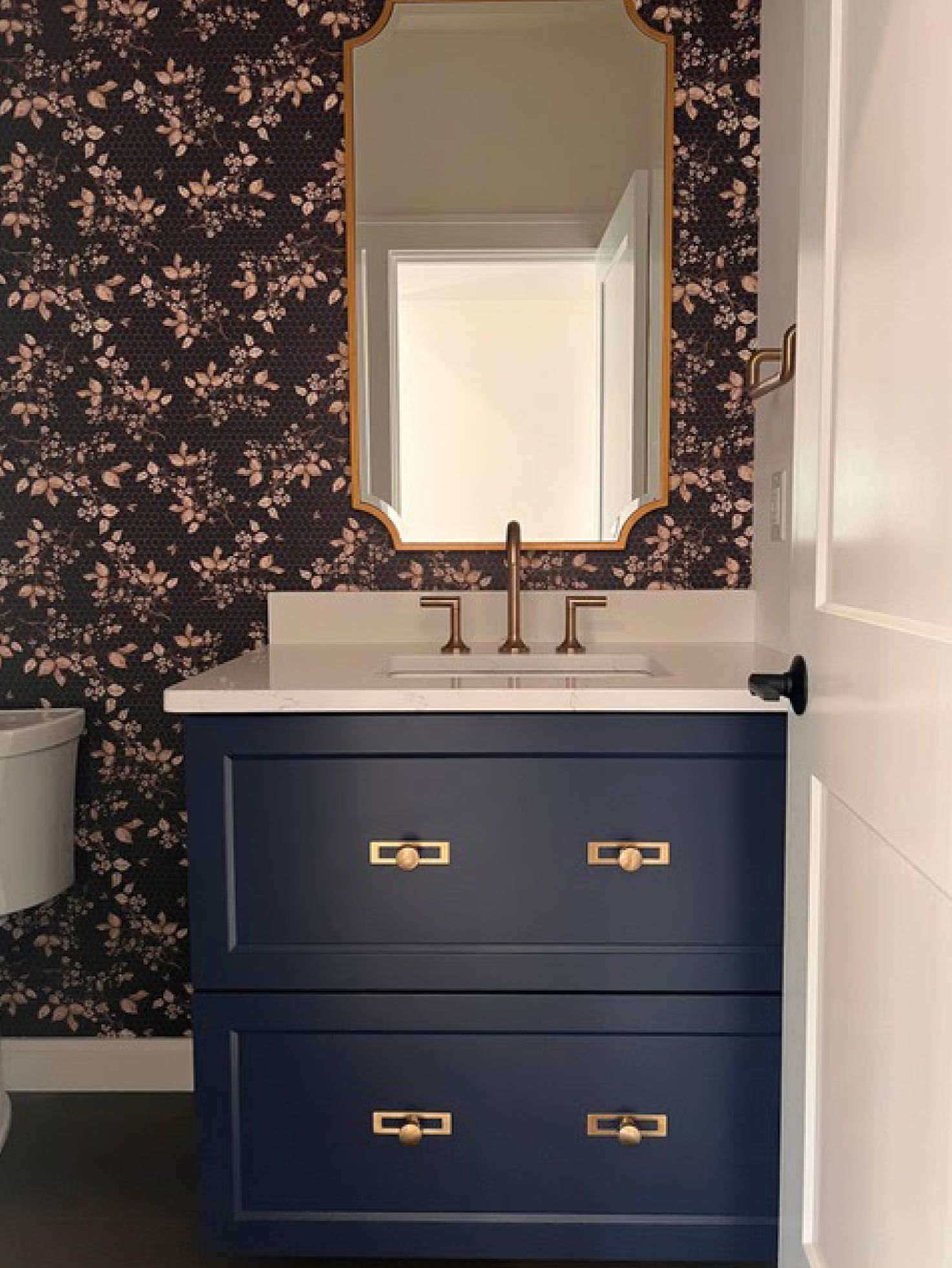 Navy bathroom vanity with gold hardware, floral wallpaper, gold-framed mirror, and white sink.