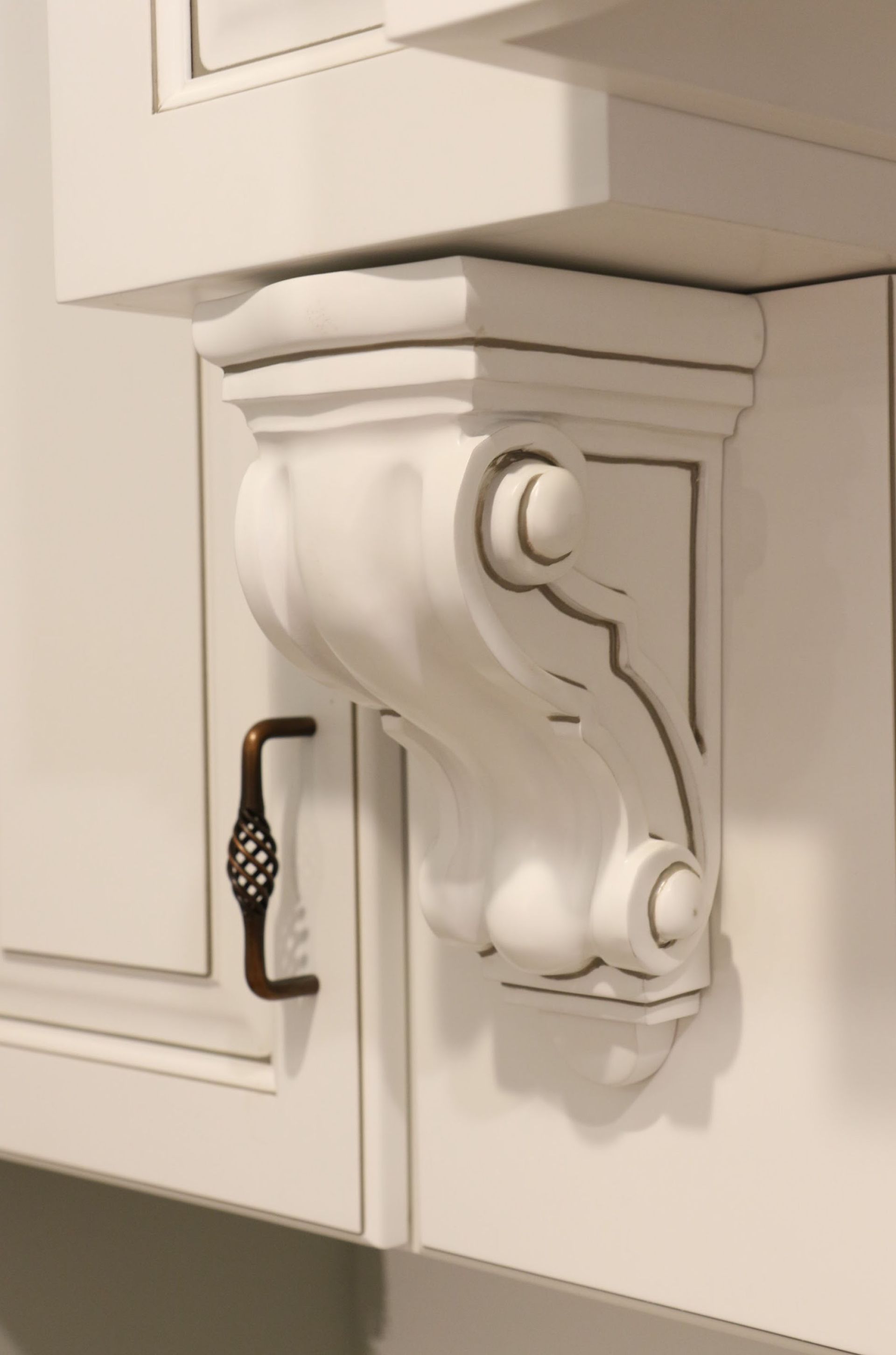 White, ornate wooden corbel supporting a kitchen cabinet, with bronze cabinet pull.