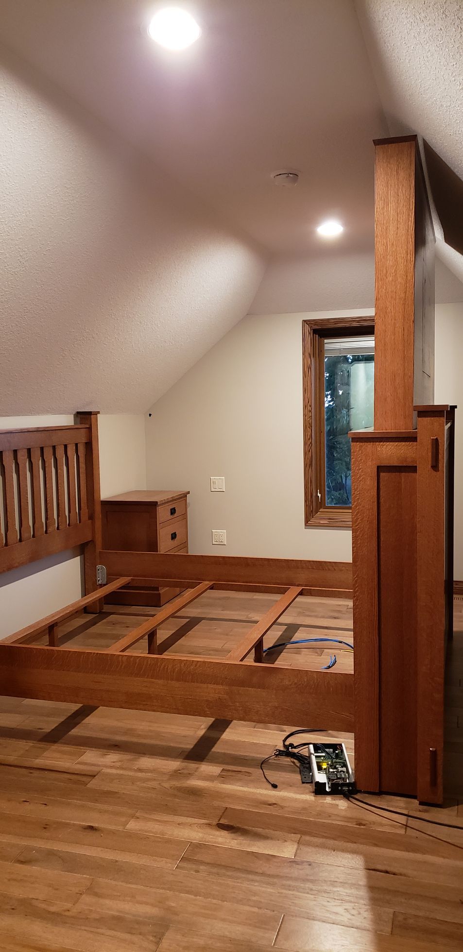A wooden bed frame and nightstand in a bedroom with sloped ceilings and a window.
