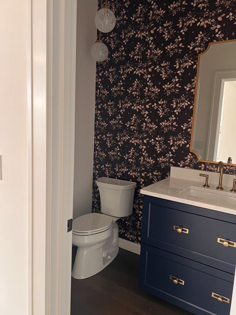 A navy blue bathroom with floral wallpaper, a white sink, toilet, and a gold-framed mirror.