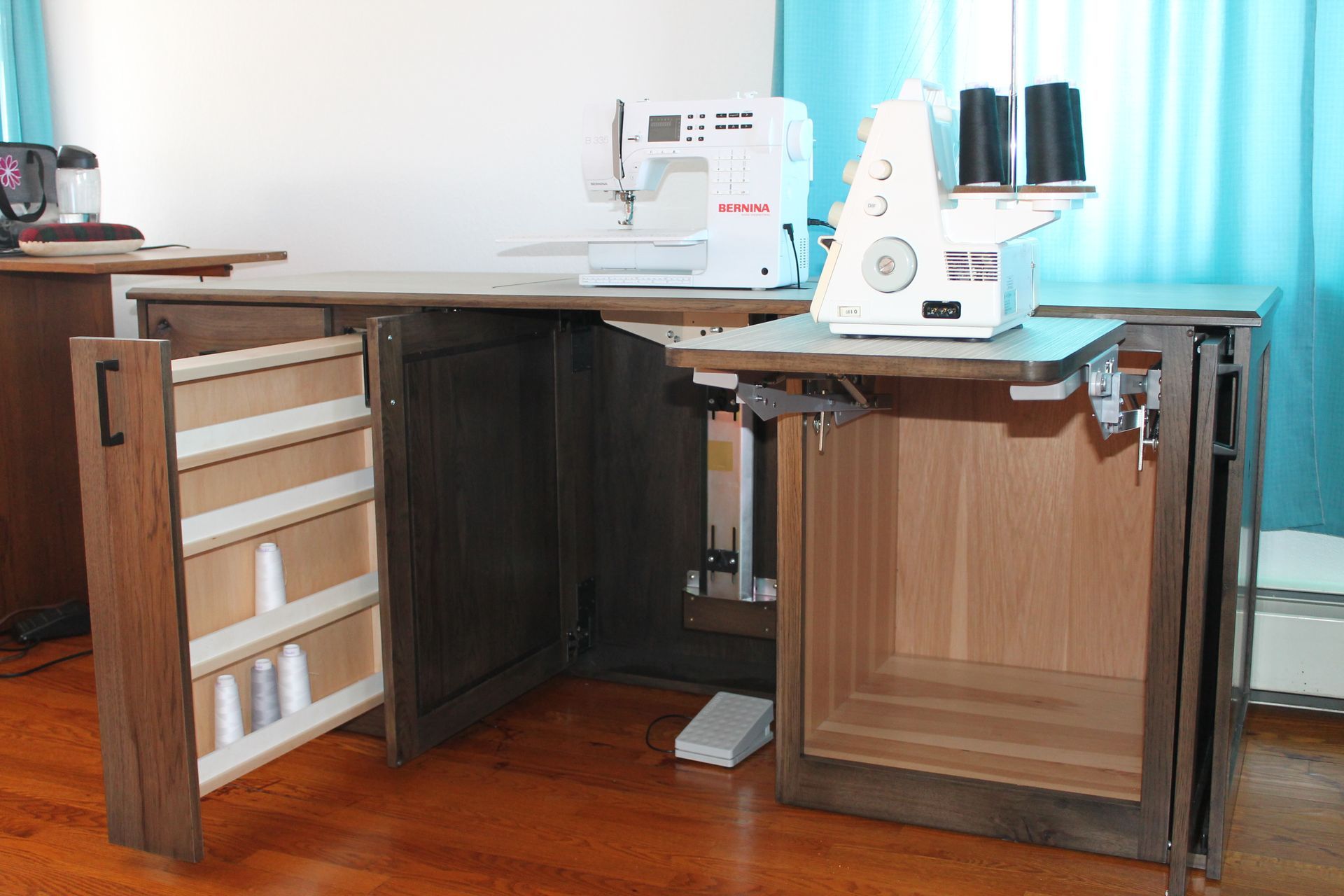 Sewing table with a machine, serger, and storage. Open cabinet with thread spools. Wooden floors and walls.