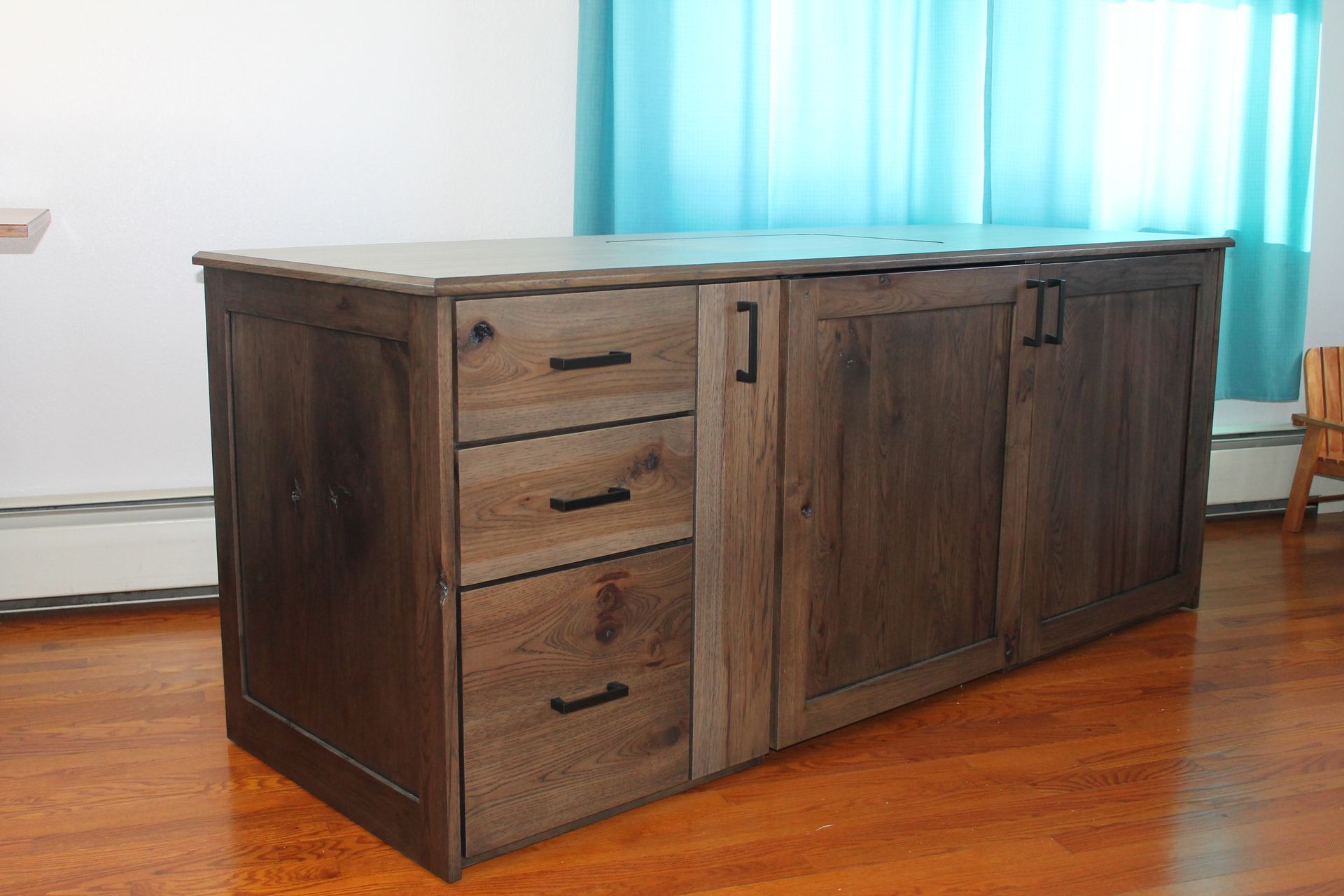 Wooden cabinet with drawers and doors, brown color. Stands on a wooden floor in front of a blue curtain.