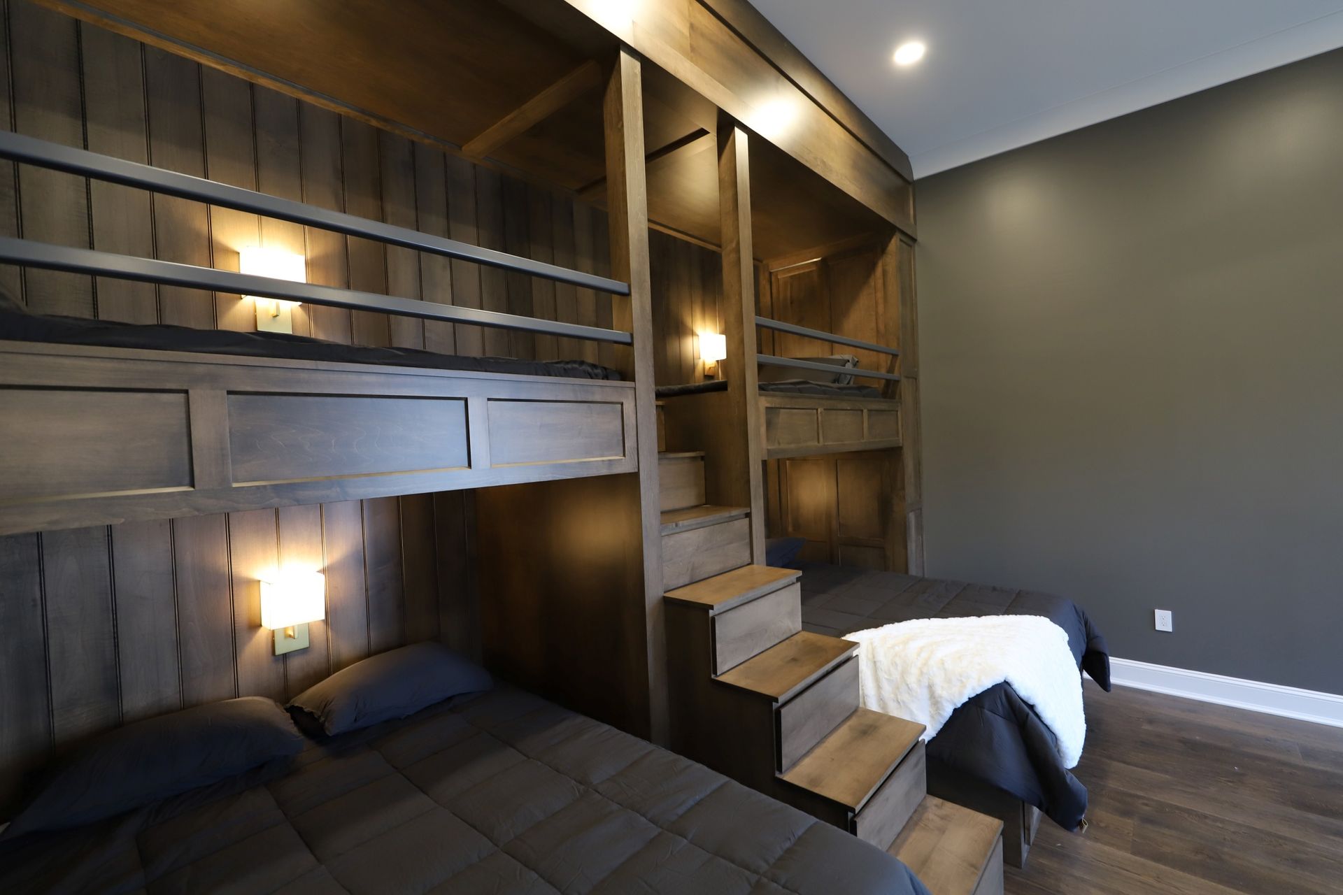 Wooden bunk beds in a room, two beds below, two above, steps leading to upper bunks, neutral tones.