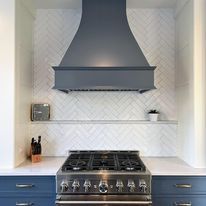 Kitchen with gray range hood, stove, blue cabinets, and white herringbone tile backsplash.