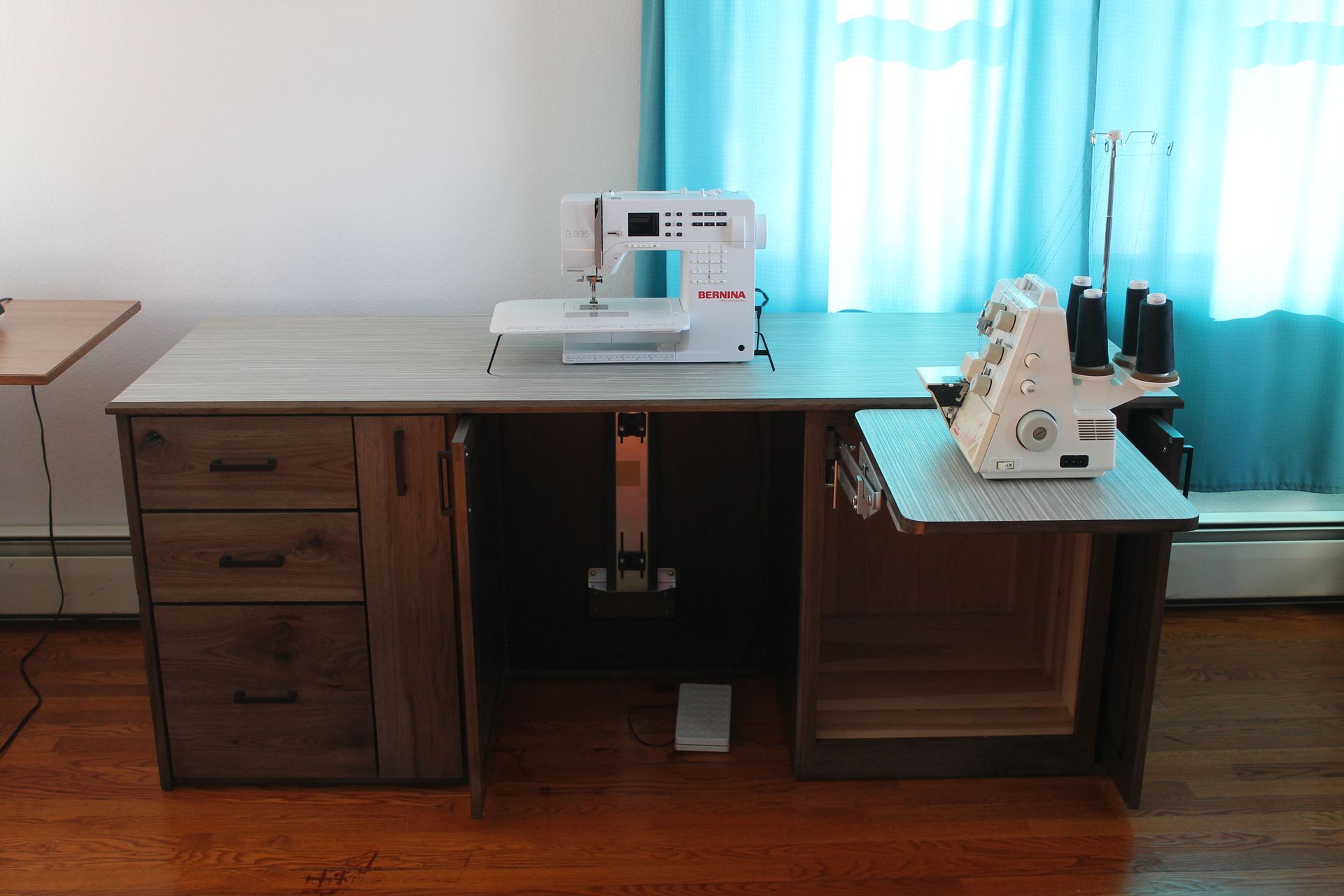 Sewing table with sewing machine and serger, wood drawers, and a blue curtain background.