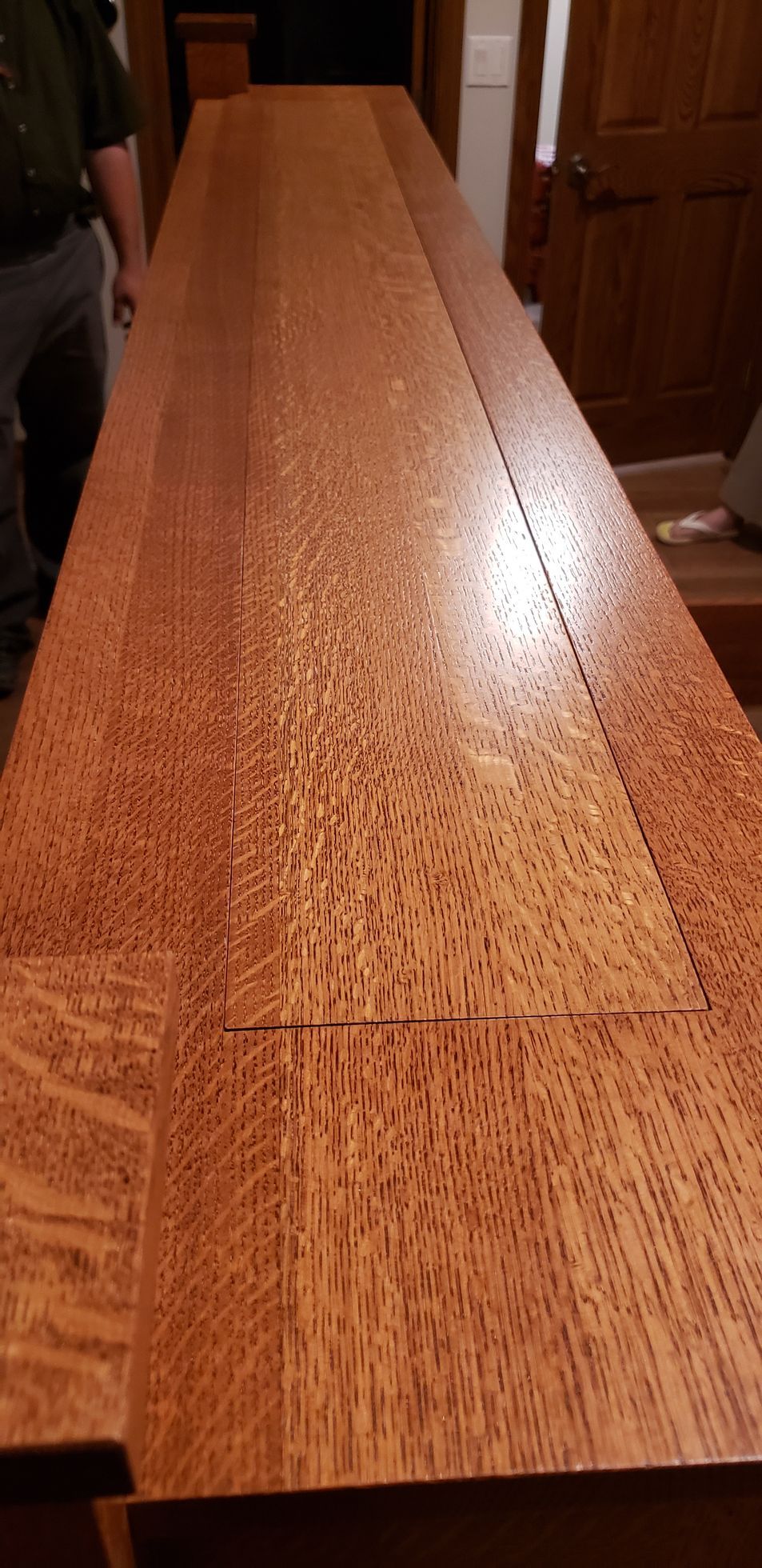 Long wooden table, with reddish-brown wood grain. A person stands to the left. Interior hallway setting.