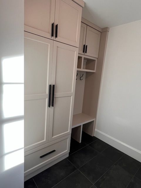 Light-colored built-in storage unit with cabinets, drawers, hooks, and a bench in a room with dark flooring.