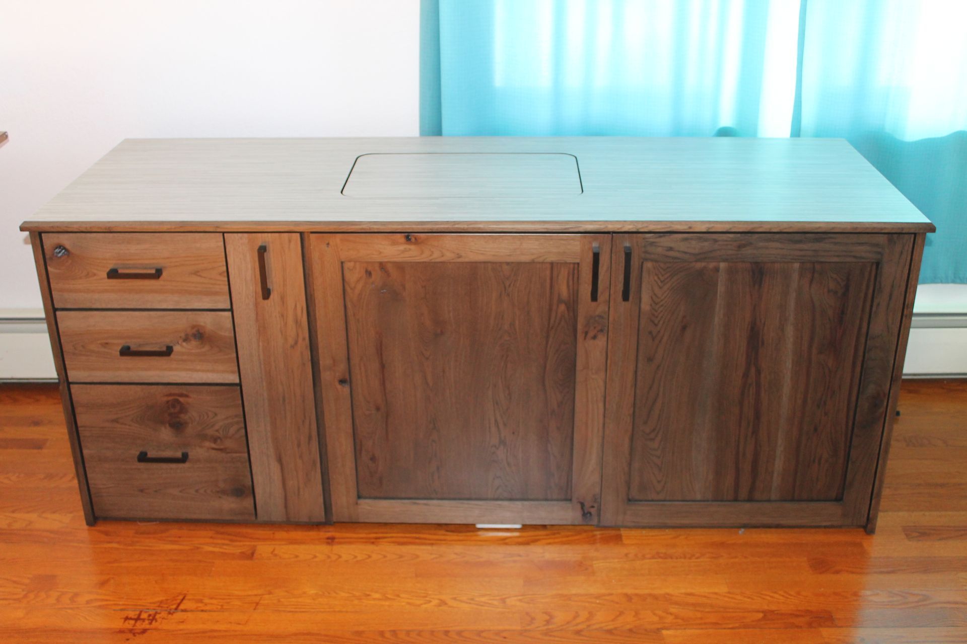 Wooden cabinet with drawers and doors, countertop with recessed space, hardwood floor, and light blue curtain in the background.