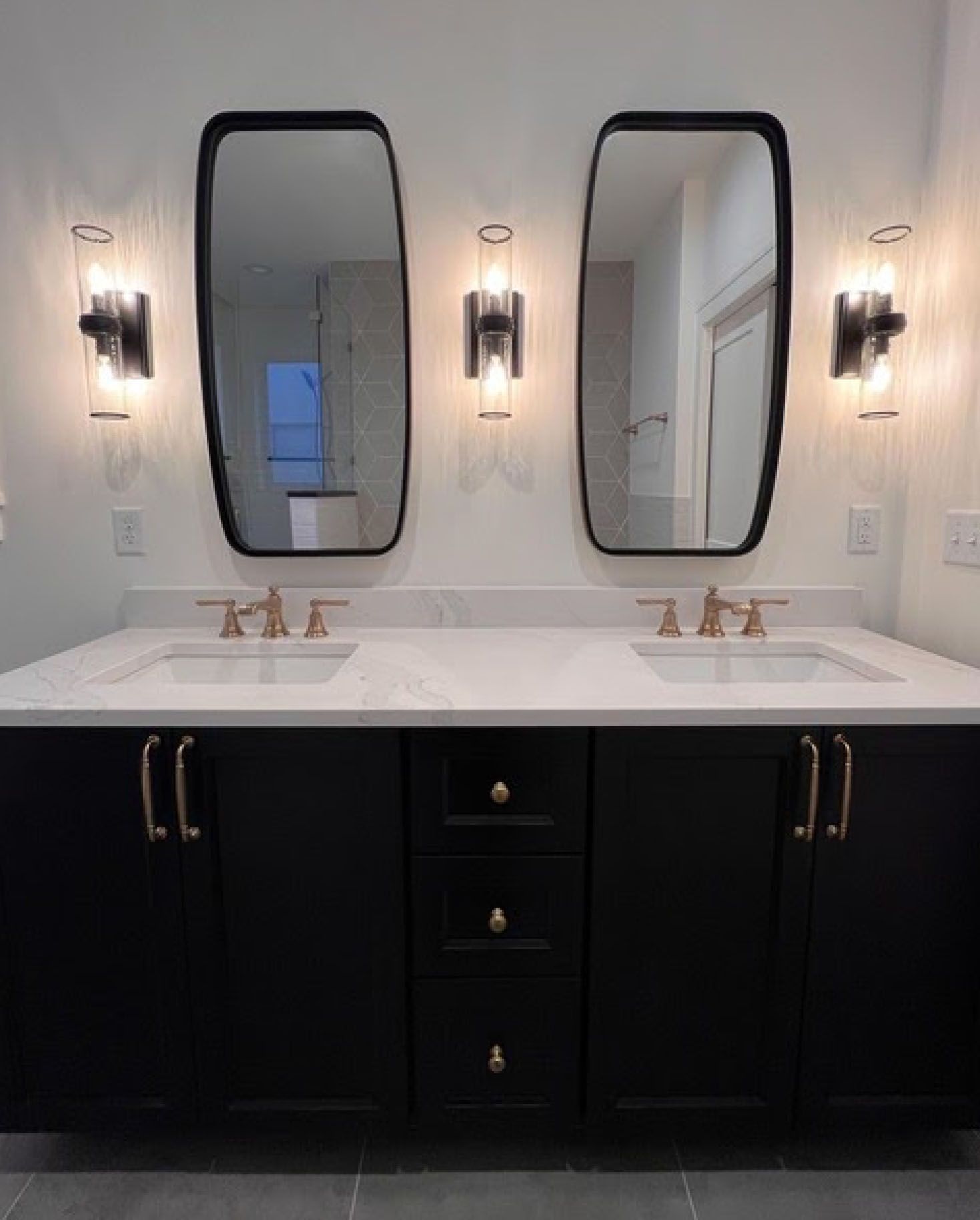 Black bathroom vanity with gold hardware, two sinks, mirrors, and sconces against a white wall.