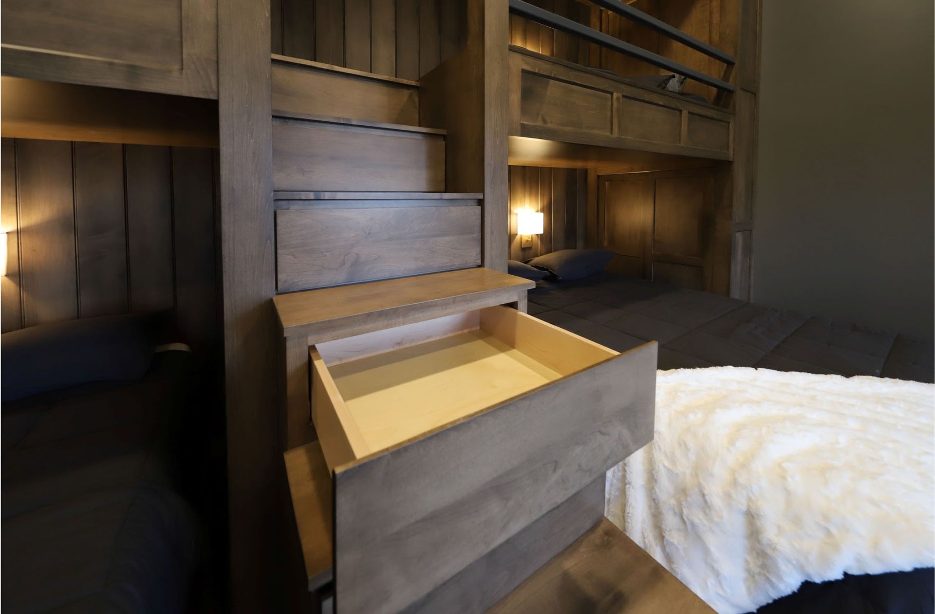 Wooden bunk beds with integrated stairs and drawers, lit by bedside lamps.