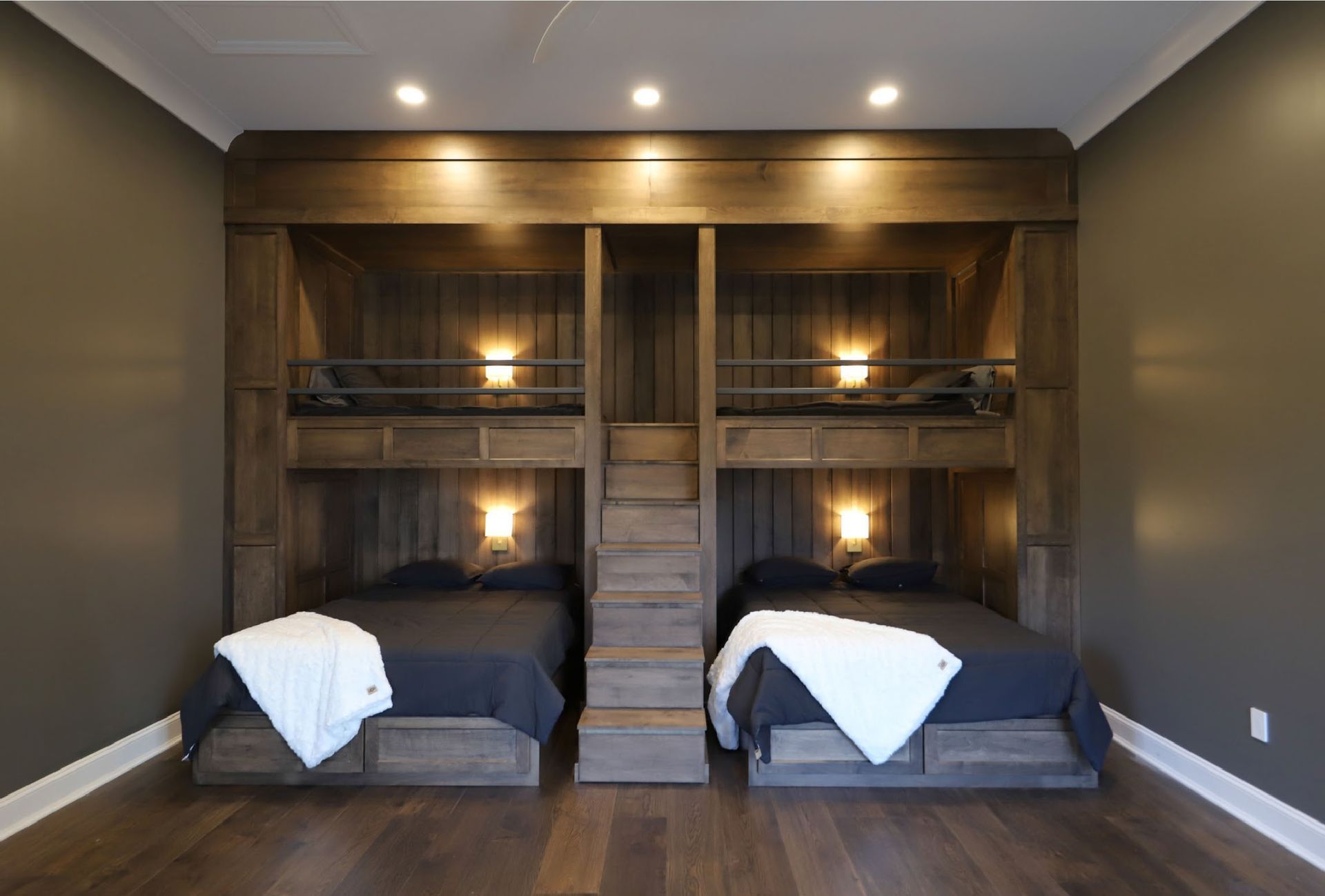 Bunk beds built into wall with upper and lower sleeping areas, wood steps, and sconce lighting.