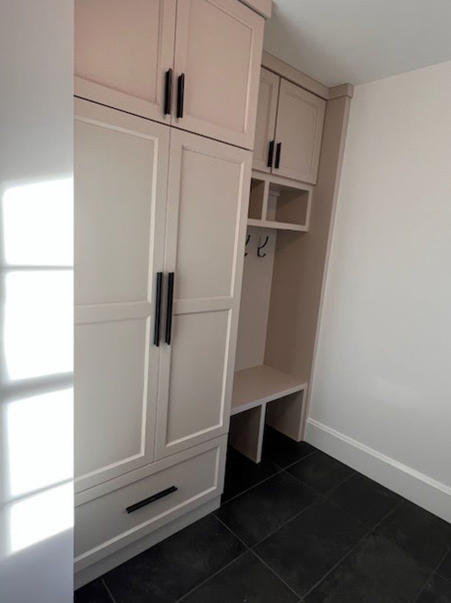 Built-in cream-colored storage cabinet and bench with black hardware in a room with dark tile flooring.