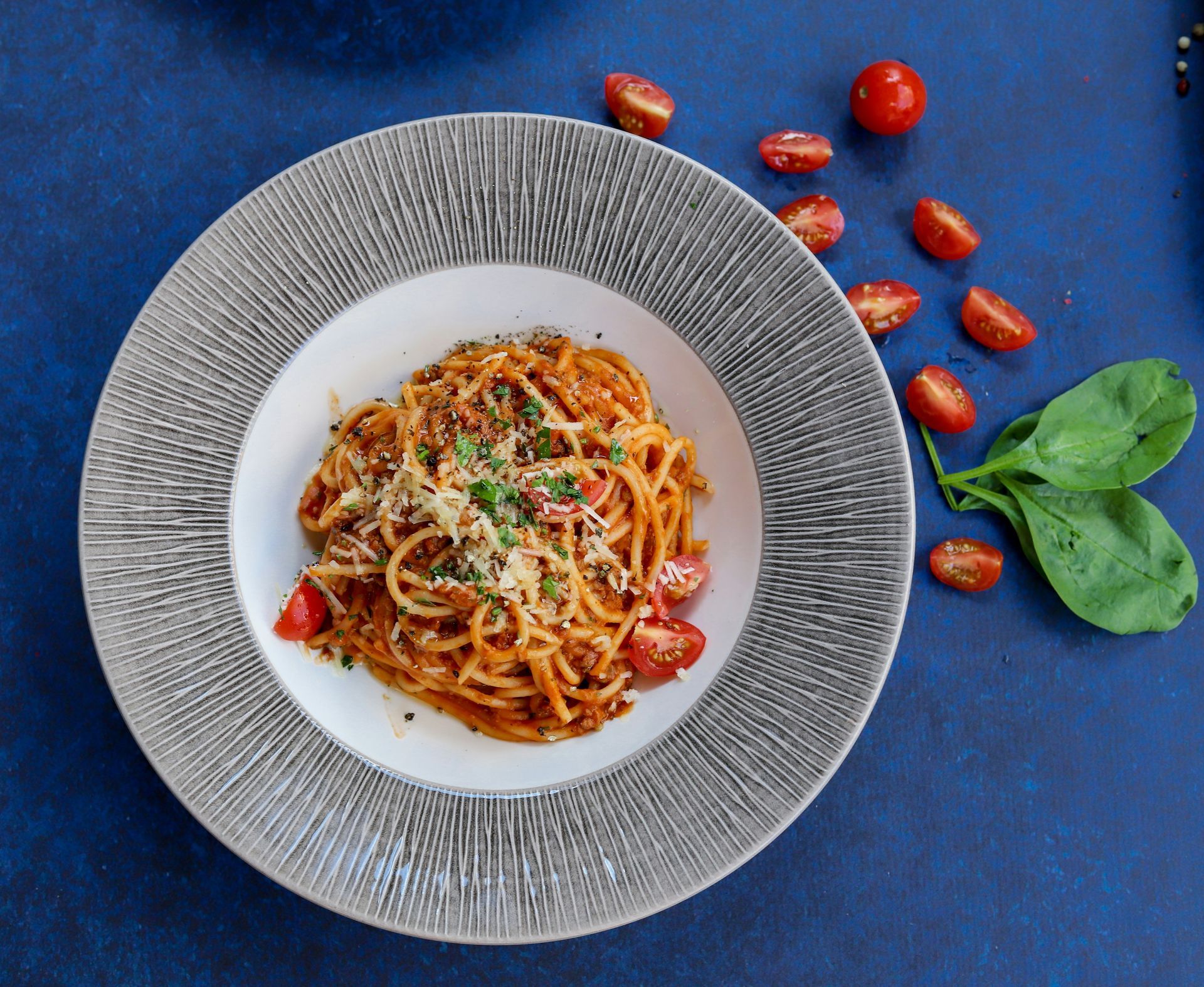Spaghetti with tomato sauce, garnished with tomatoes and parsley, on a textured plate, beside spinach and halved tomatoes.