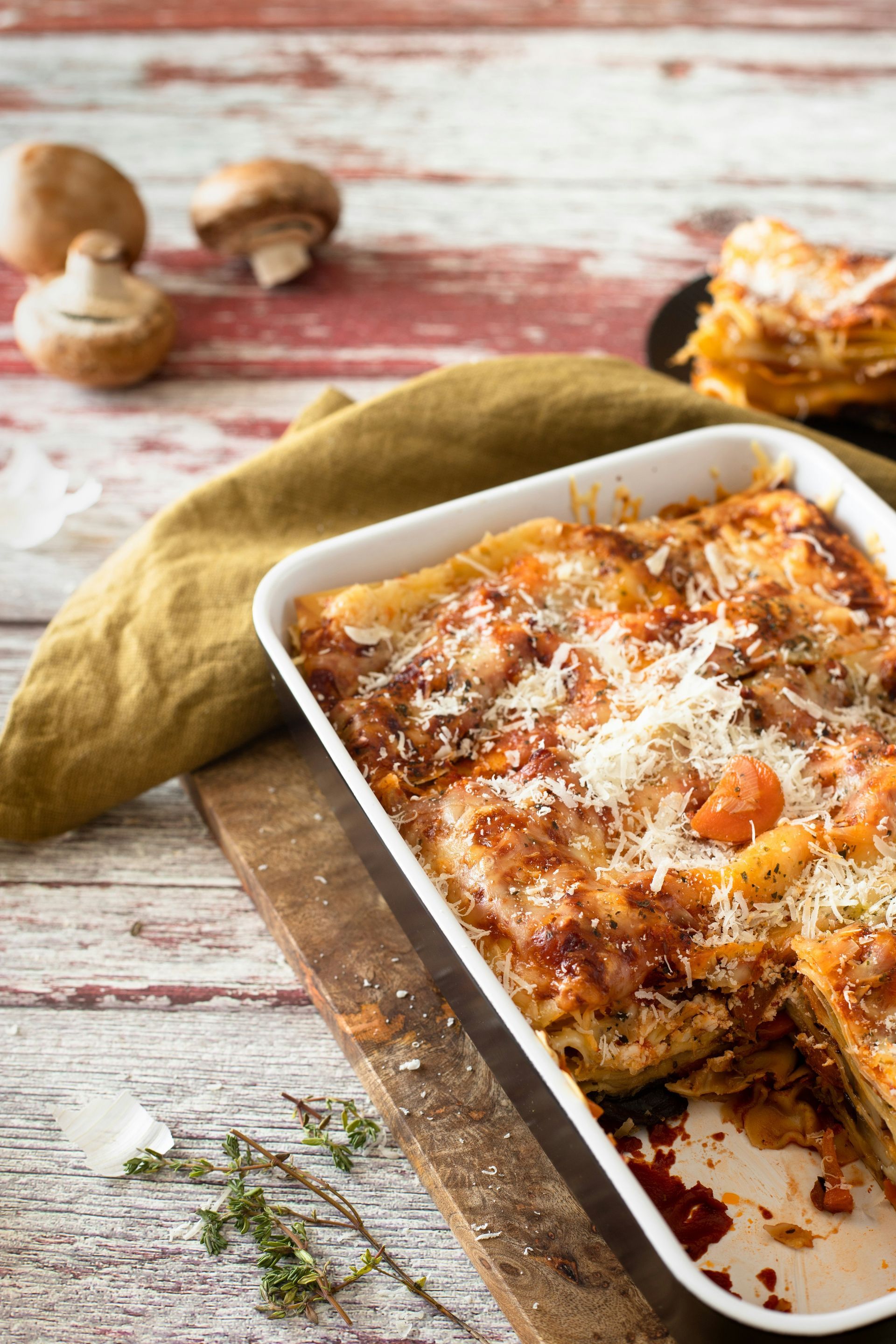 Lasagna in a white dish, on a wooden board, next to mushrooms and a green cloth.