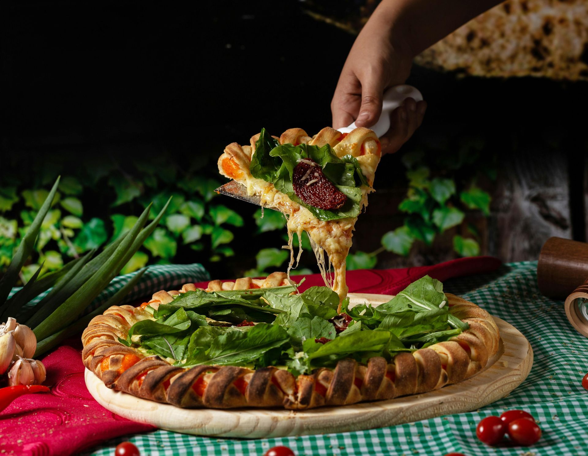 Hand lifting a slice of pizza with melted cheese, arugula, and sun-dried tomatoes. Pizza on a wooden board, green leafy background.