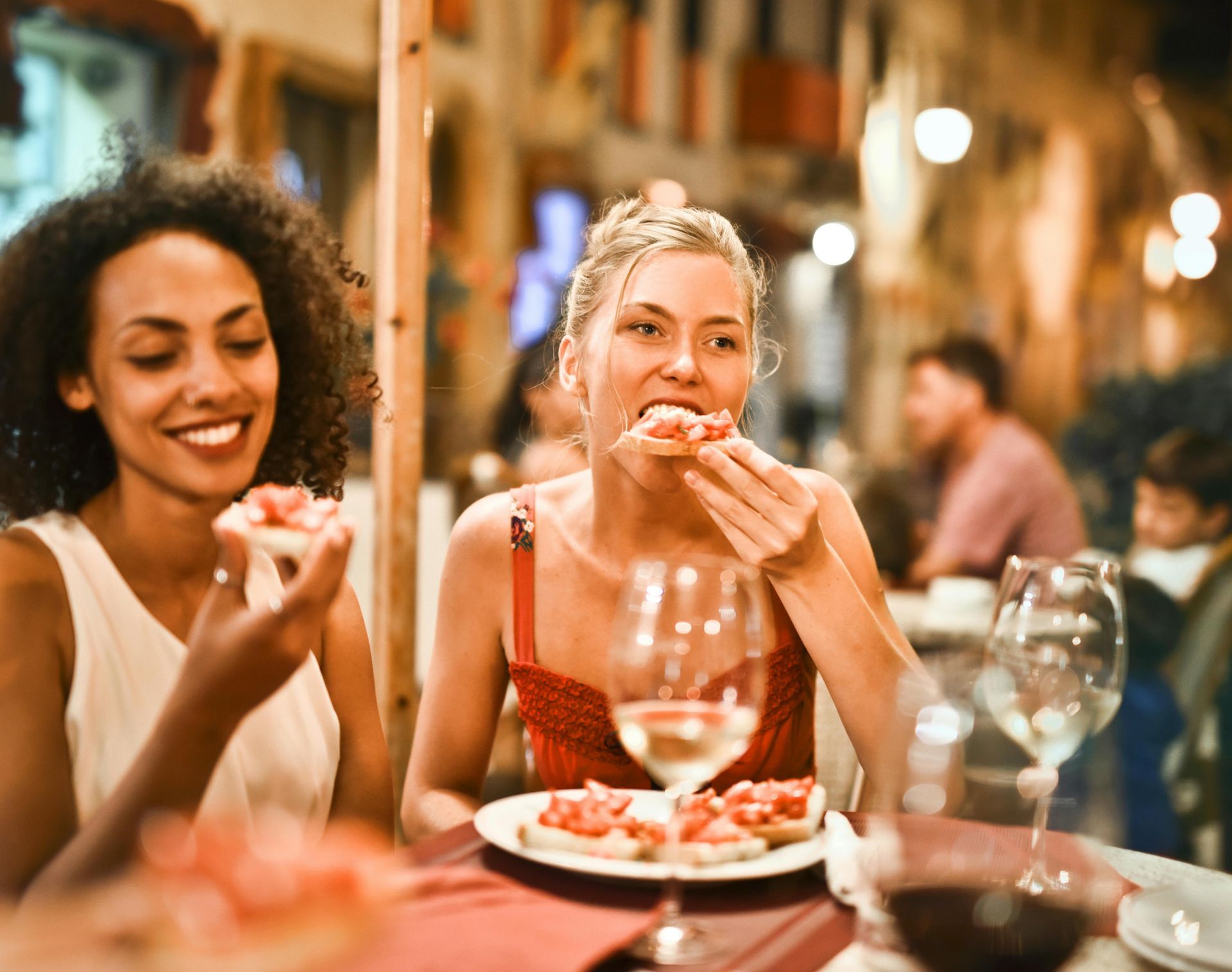 Two women eating bruschetta at a restaurant, smiling. One woman wears a red dress, the other, white.