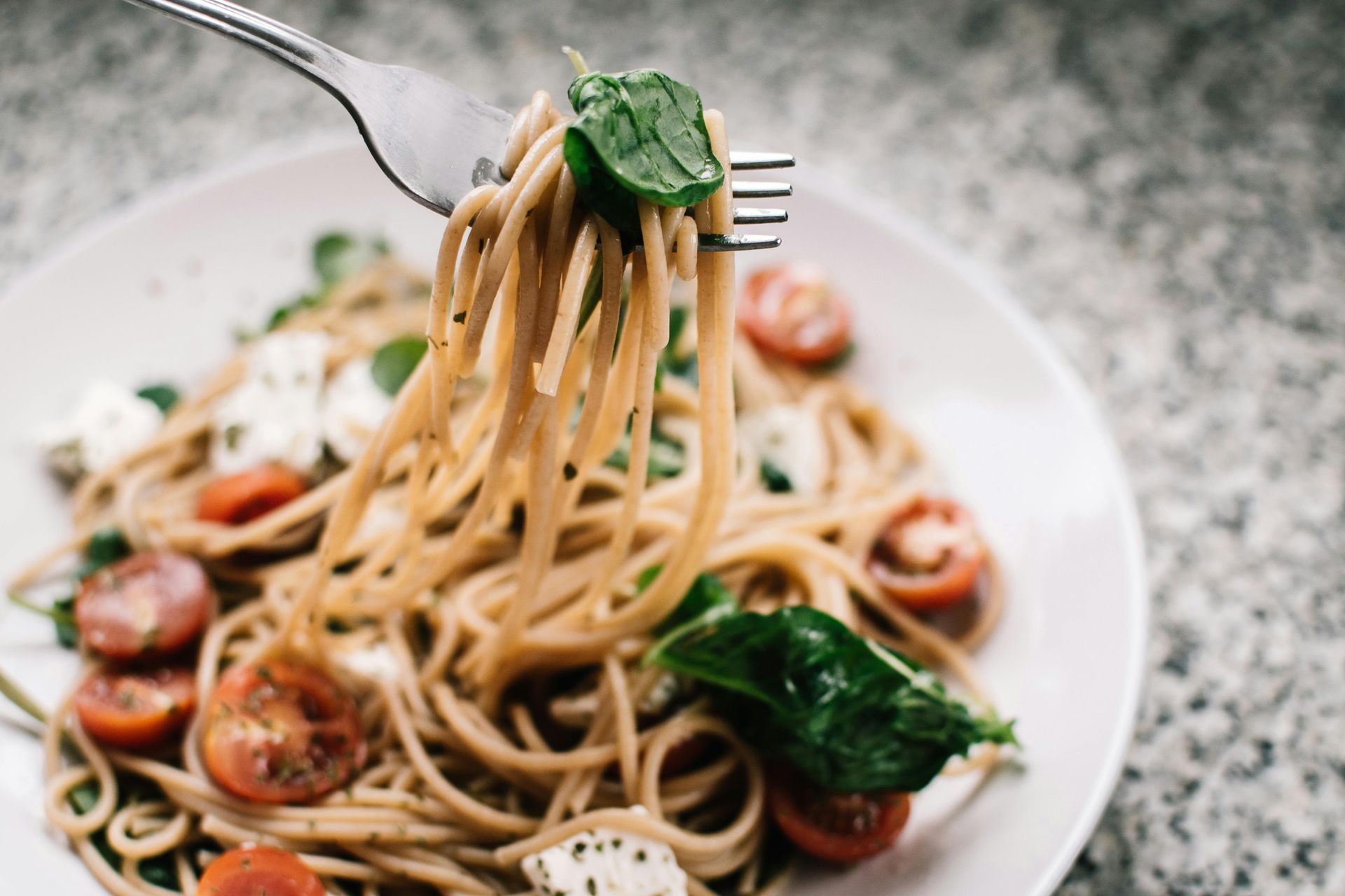 Forkful of spaghetti with tomatoes, basil, and mozzarella in a white bowl on a countertop.