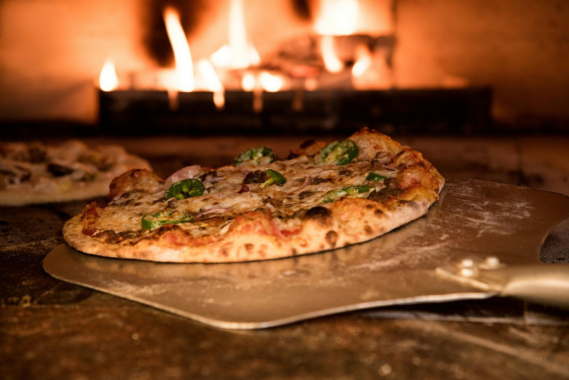 Pizza on a metal peel, inside a wood-fired oven, with flames in the background.