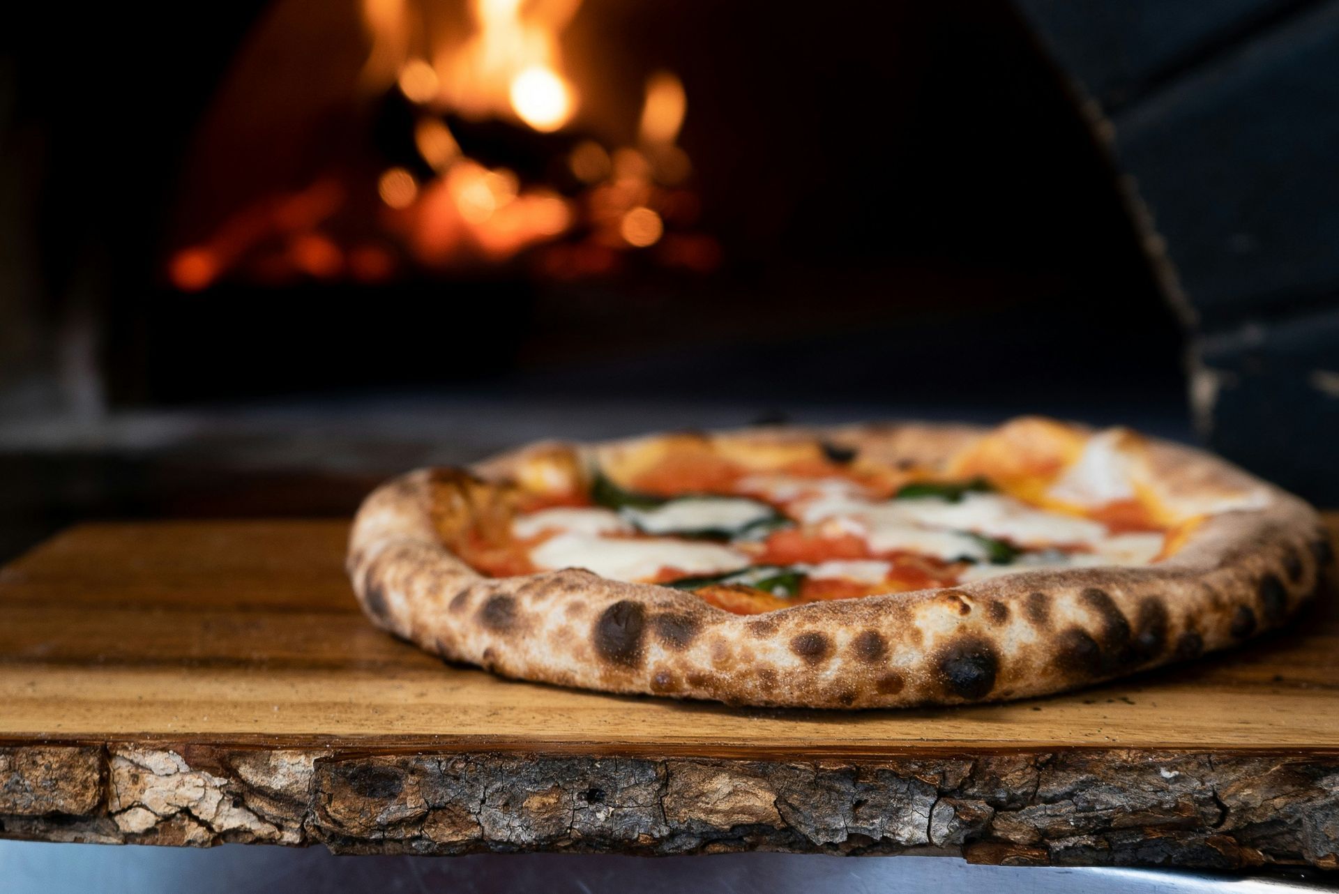 Pizza on wooden board in front of a wood-fired oven with flames.