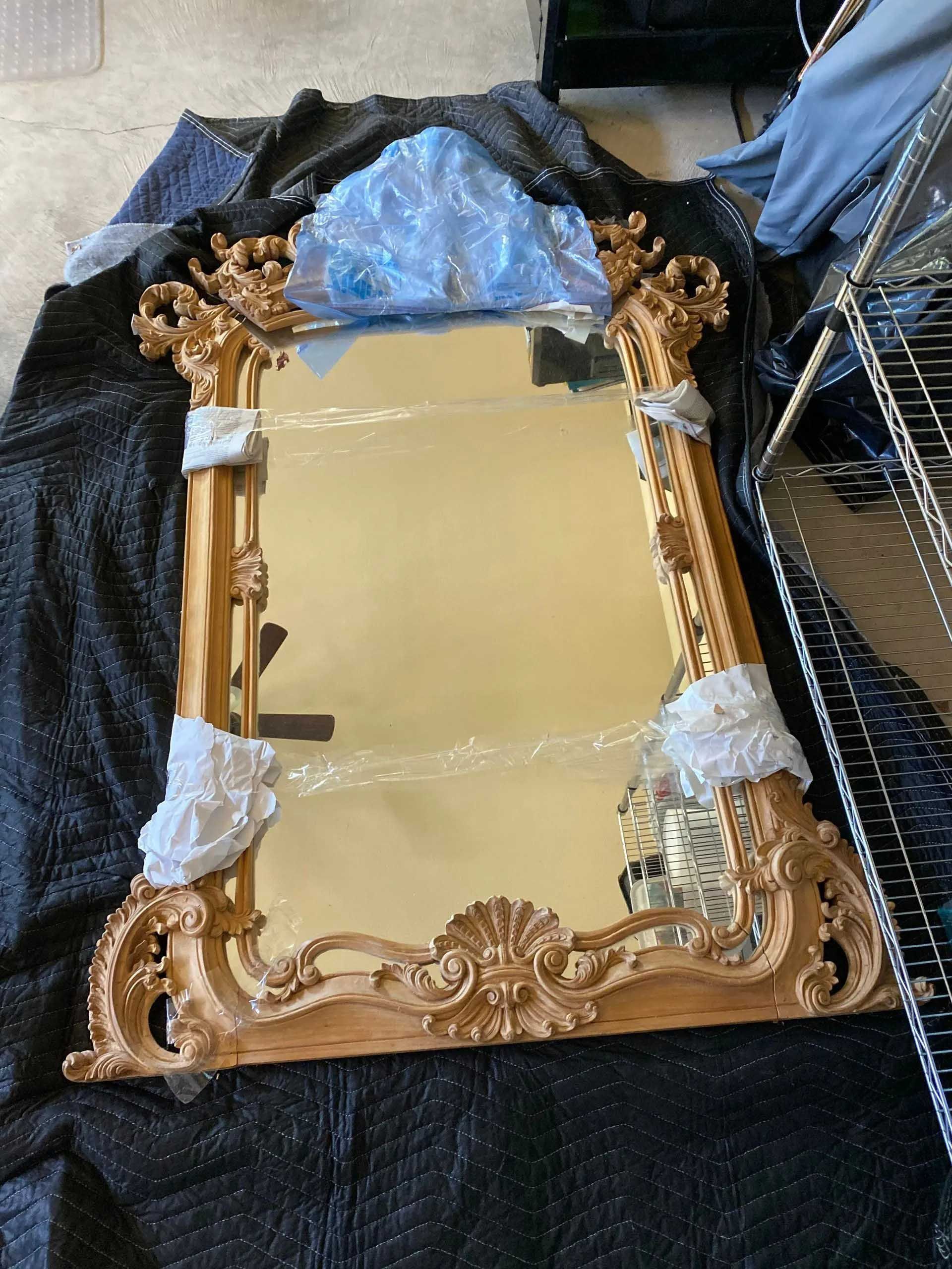Ornate gold framed mirror, partially wrapped in protective materials, on a black surface.