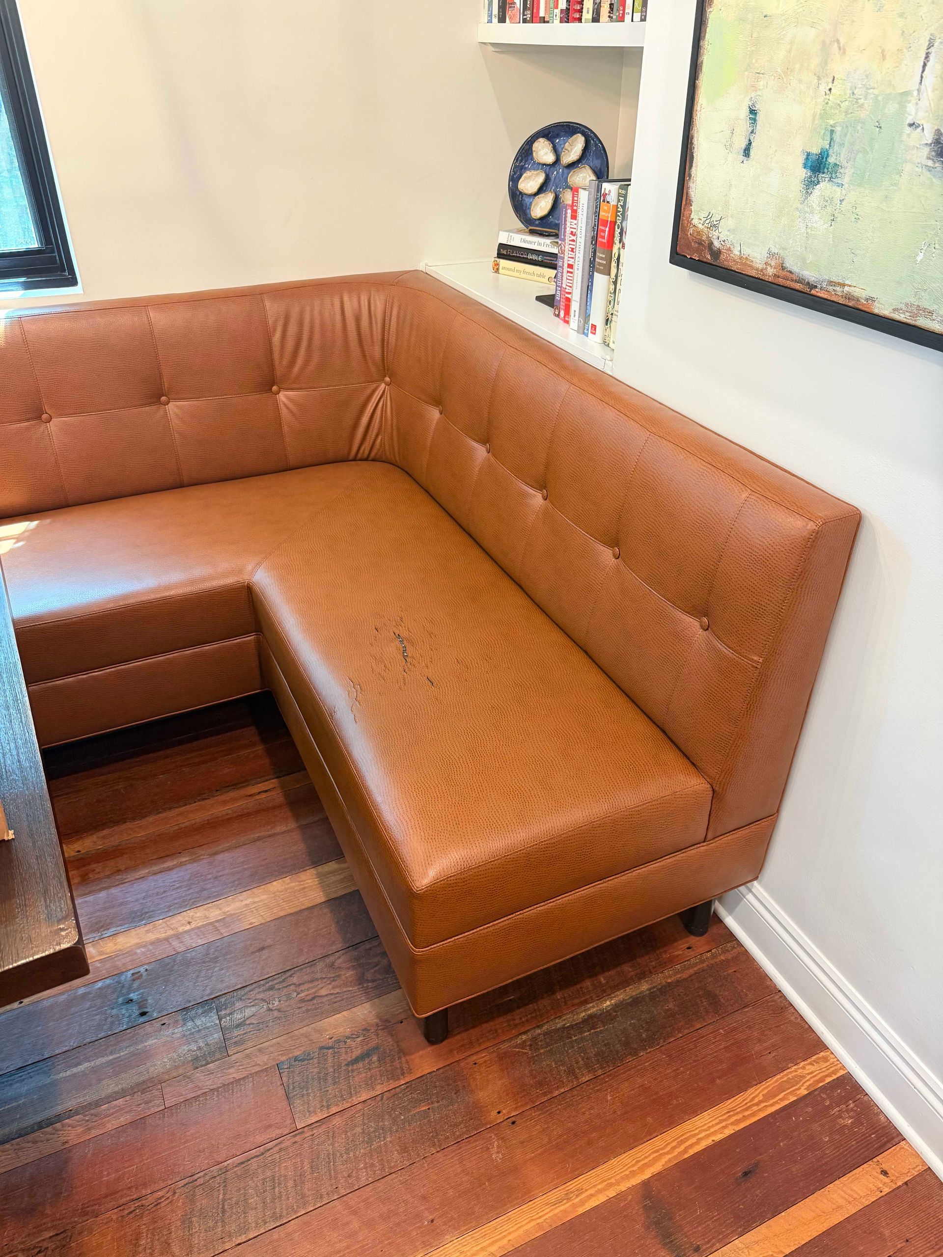 L-shaped brown leather booth in a dining nook with dark wood floor, next to a shelf and wall art.