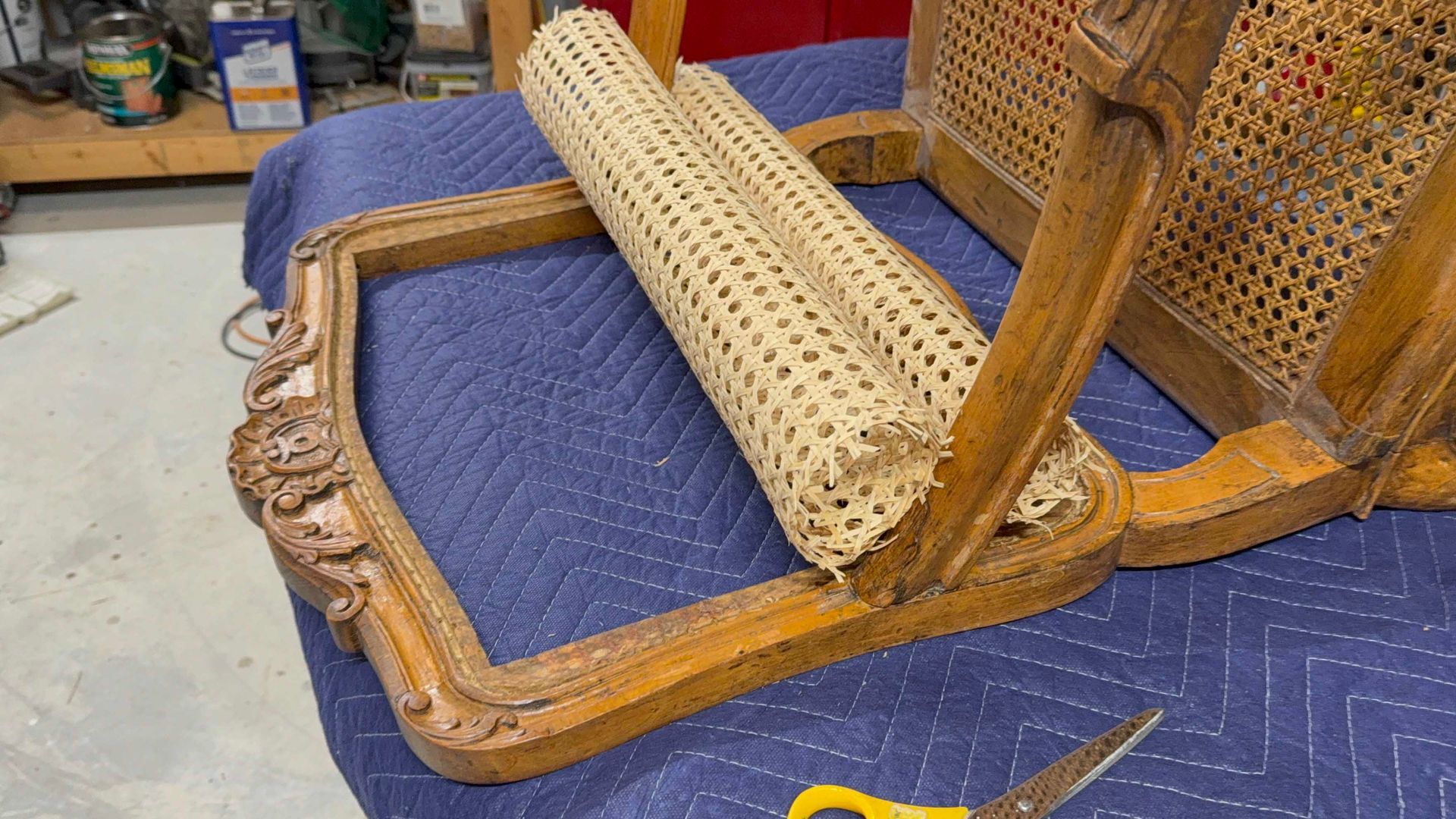 Wooden chair with broken cane seat, scissors on a blue drop cloth in workshop.