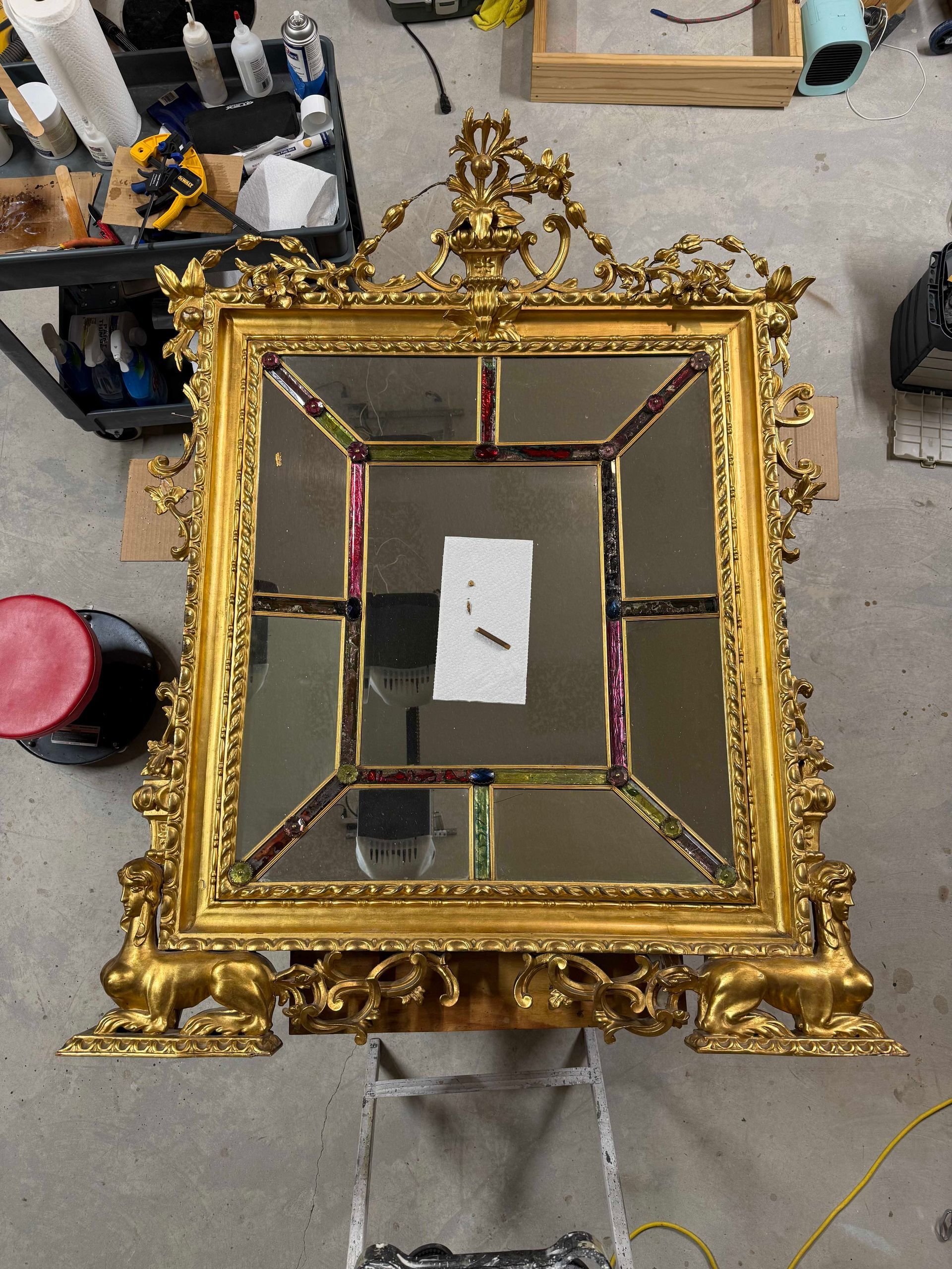 Ornate gold mirror, square design with multiple panes, resting on a silver stand, workshop background.