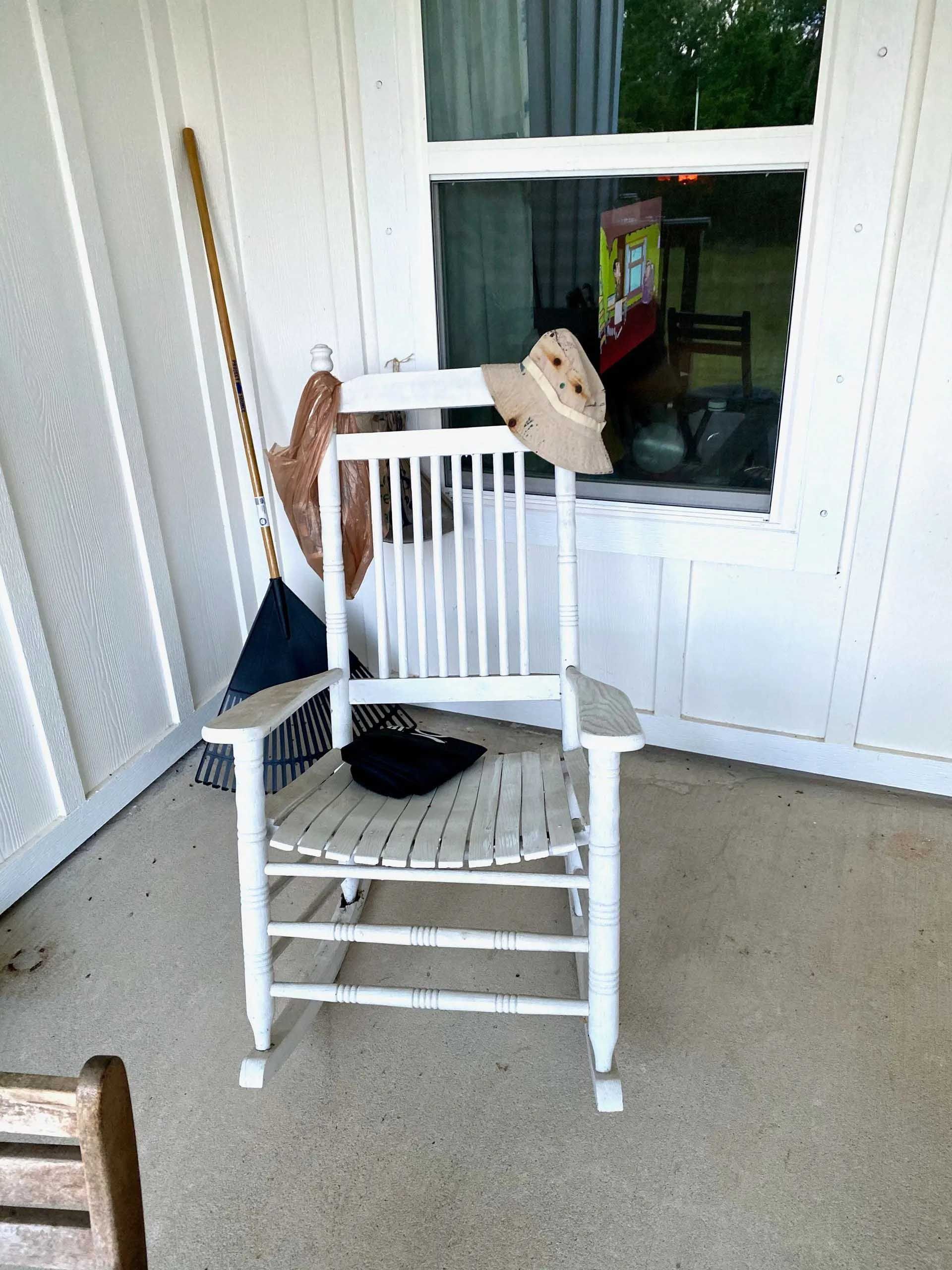 White rocking chair on a porch with a broom, hat, and window.
