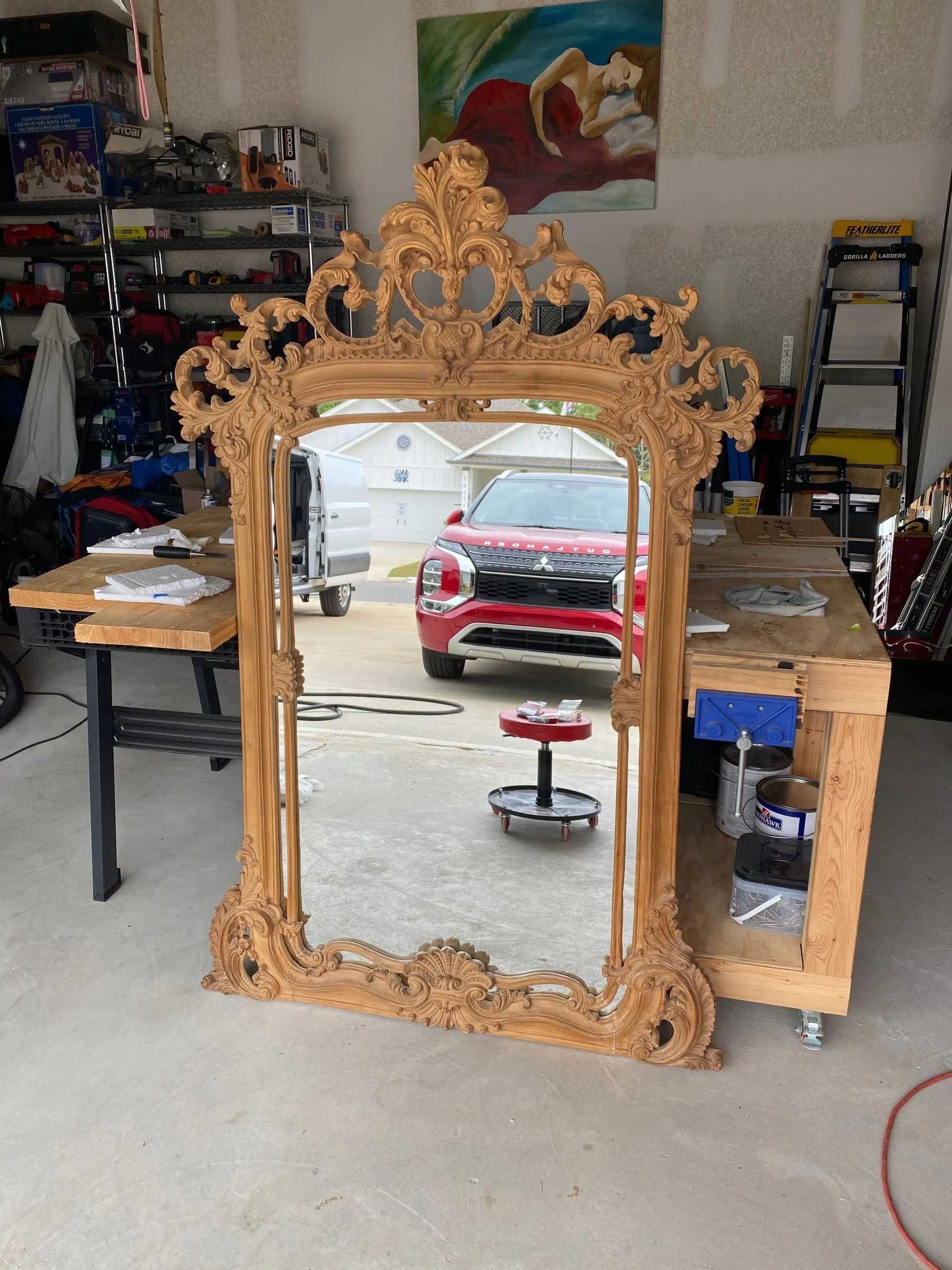Large ornate gold-framed mirror leans against a garage wall; reflection shows a red car and work area.