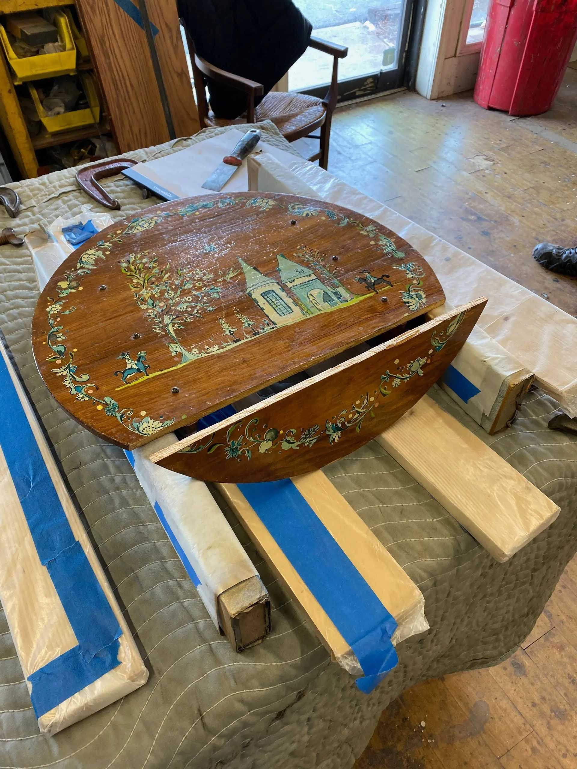 Oval wooden table top in two pieces, with painted landscape, on a workbench.
