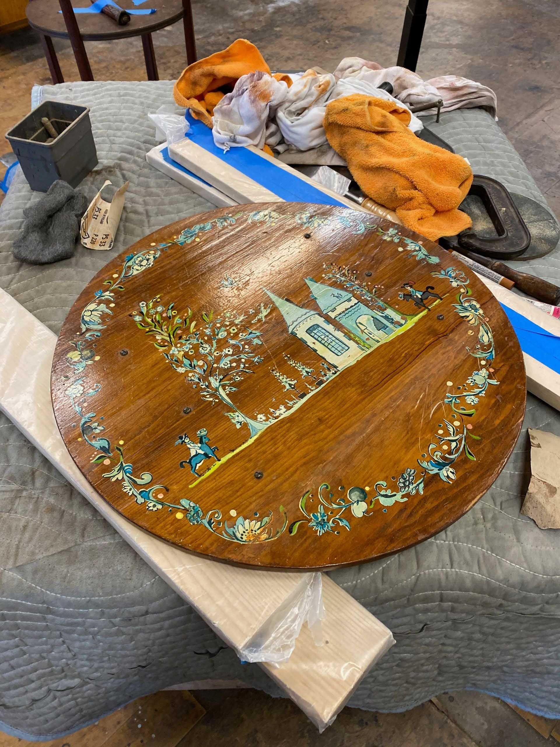 Round wooden table top with painted landscape scene and various tools on a work surface.