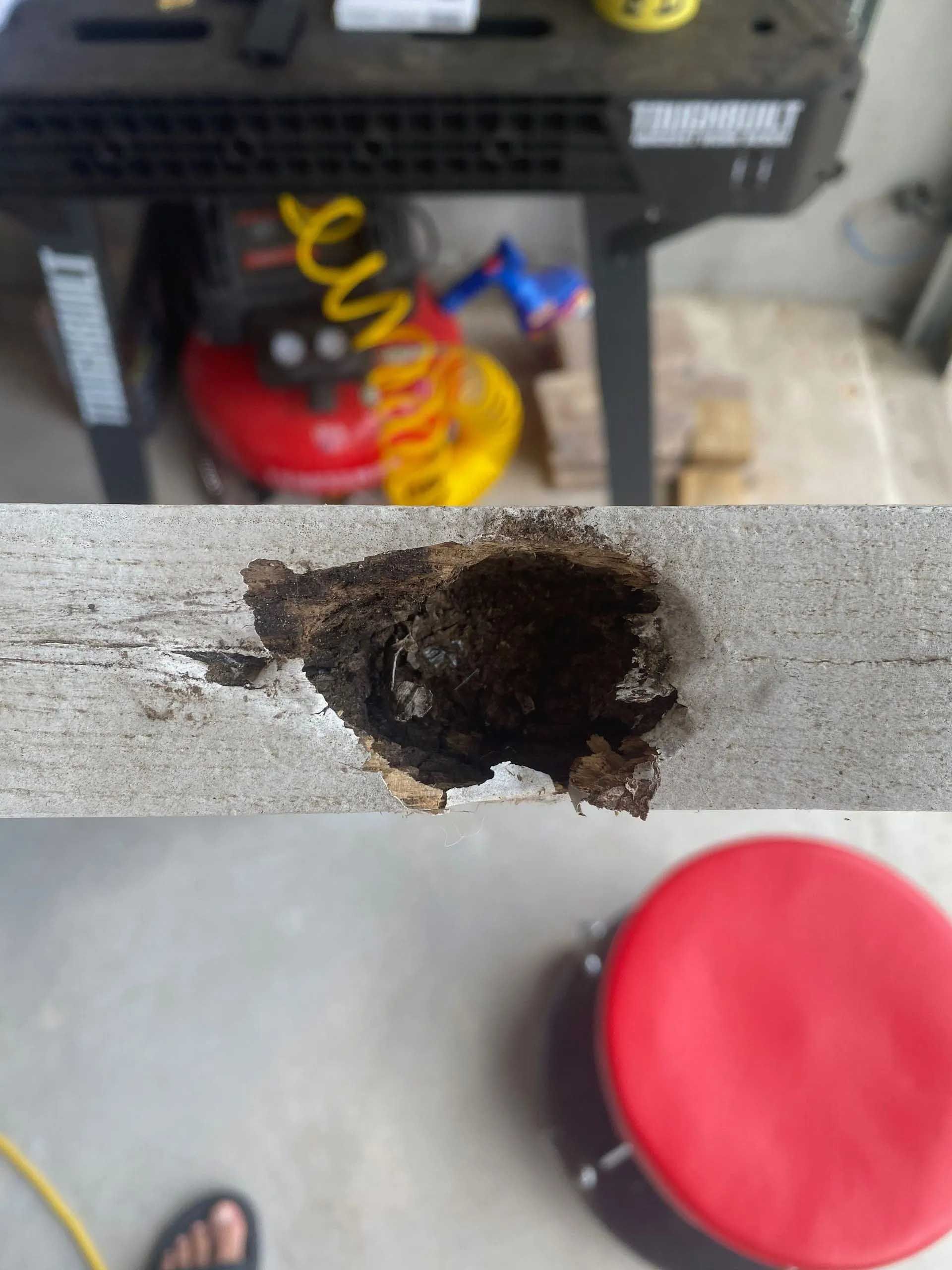 Close-up of a damaged, weathered wooden board with a large hole filled with dark debris. A red stool and tools are in the background.