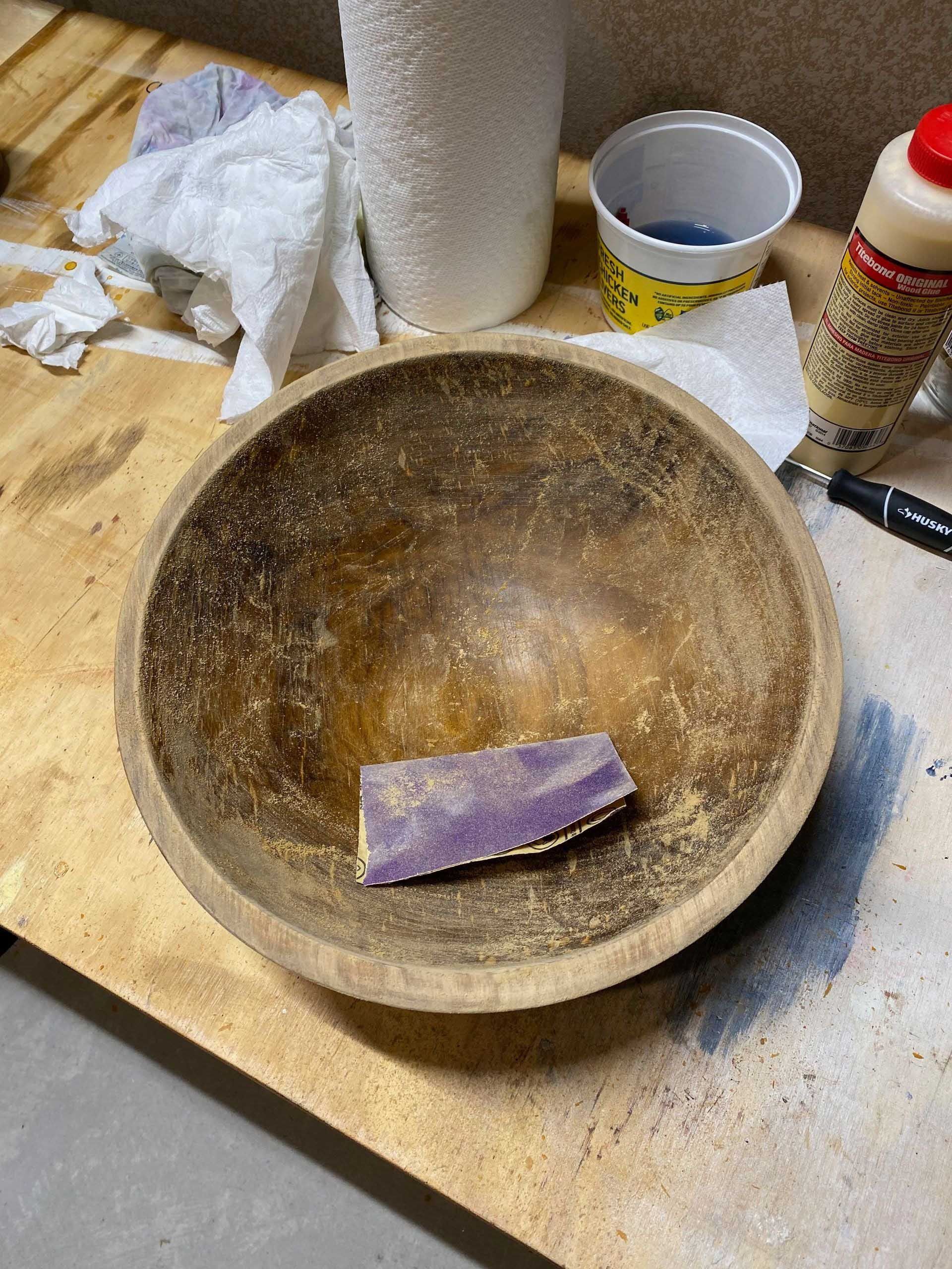 Wooden bowl with sandpaper inside, on a workbench, surrounded by supplies.