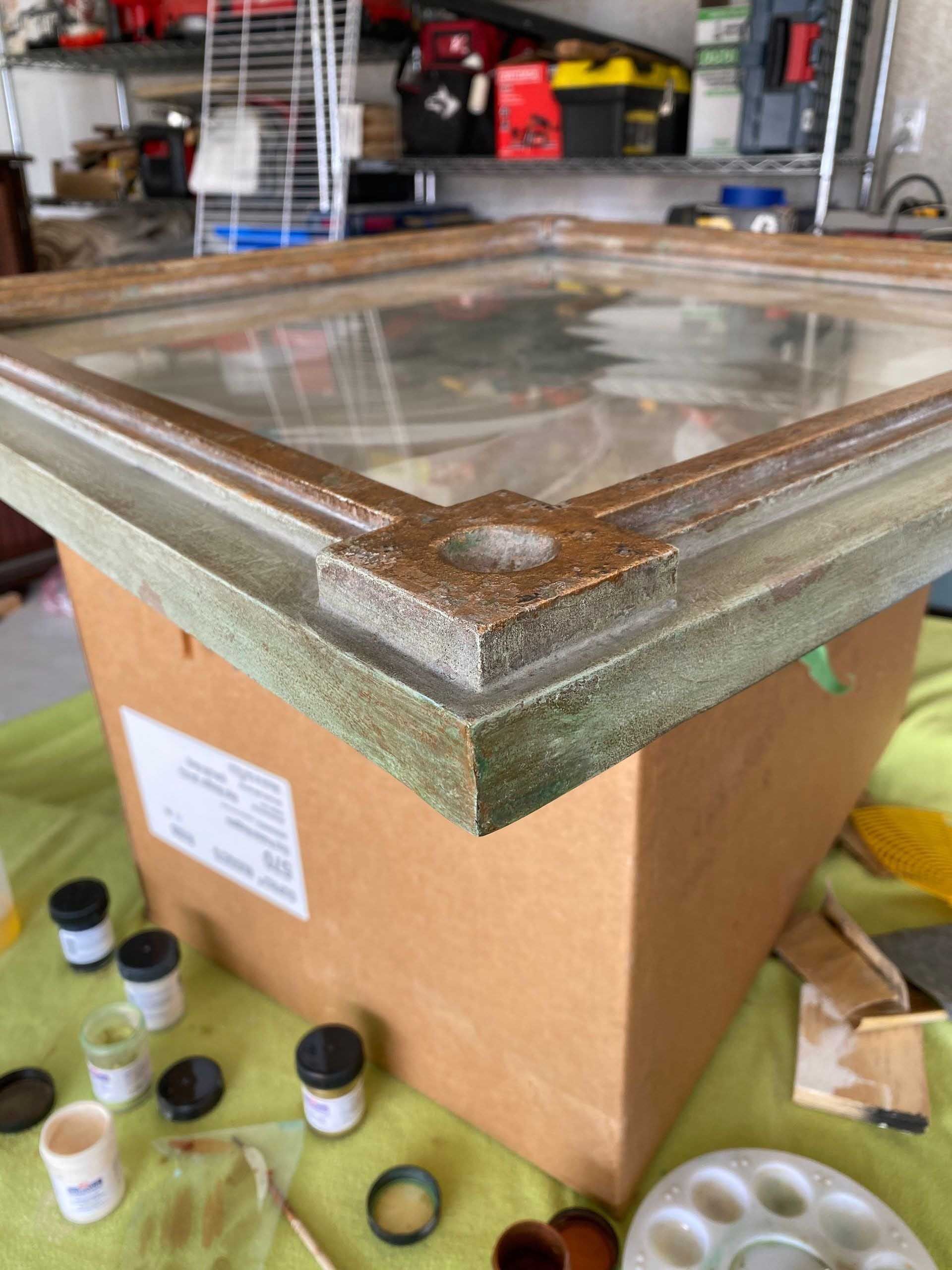 A frame with glass, being worked on, sitting on a cardboard box, with paint supplies.