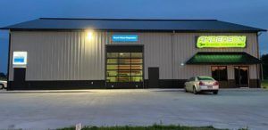 A wide commercial building at dusk with a large garage door, a green storefront sign, and a car parked in front.
