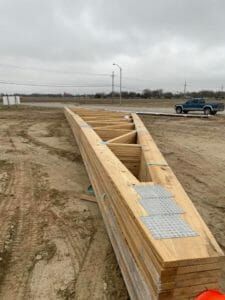 A large wooden roof truss lies flat on a dirt construction site, with a pickup truck in the distance under a cloudy sky.