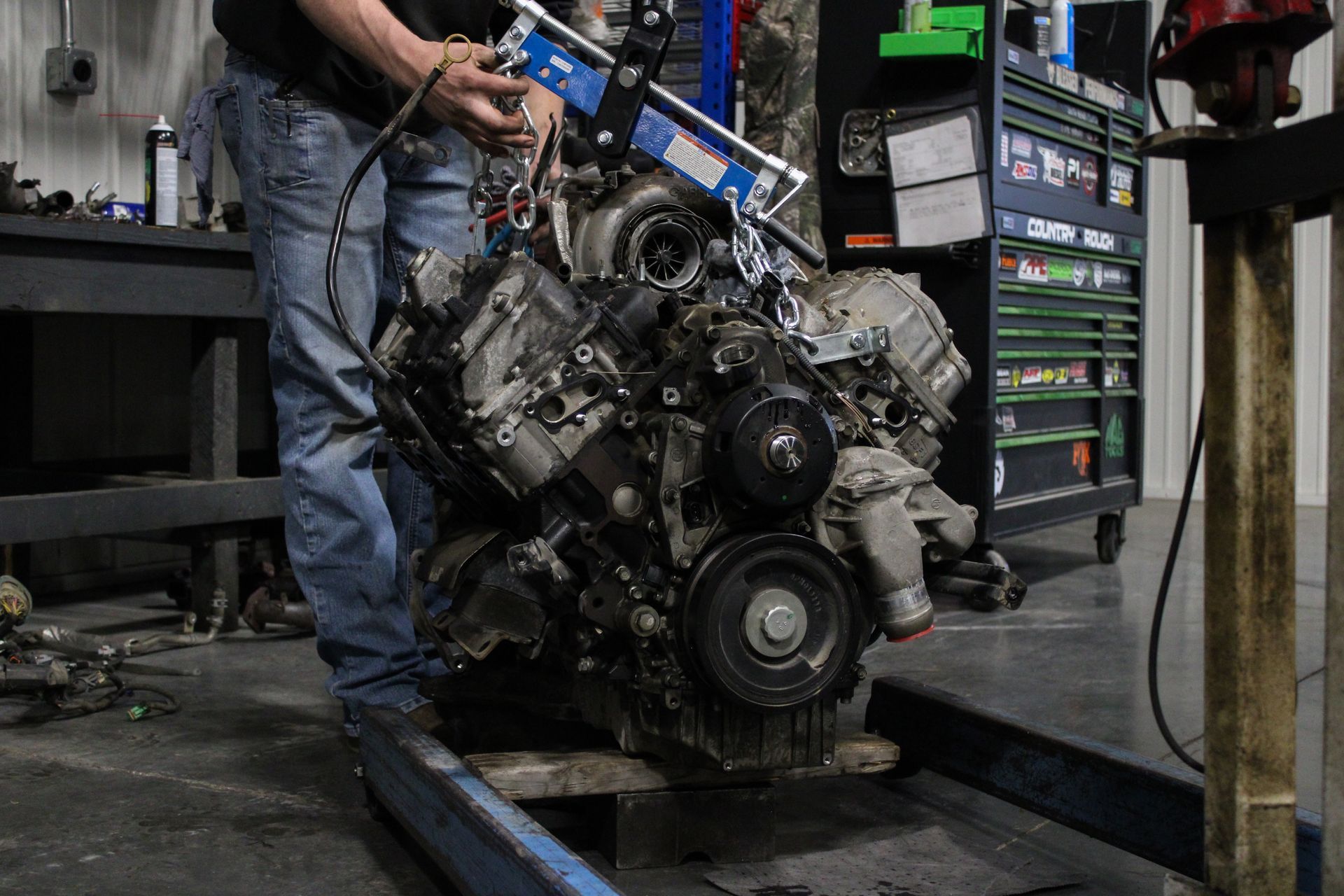 Mechanic handling a large engine block in a workshop, with tools and shop equipment nearby