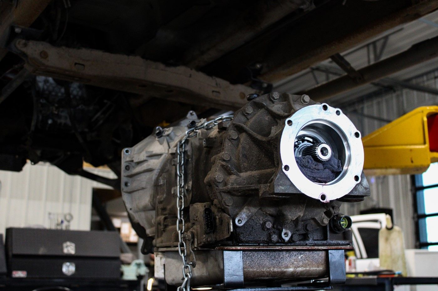 Car transmission on a stand in a workshop, with a chain hoist and yellow lift arm nearby