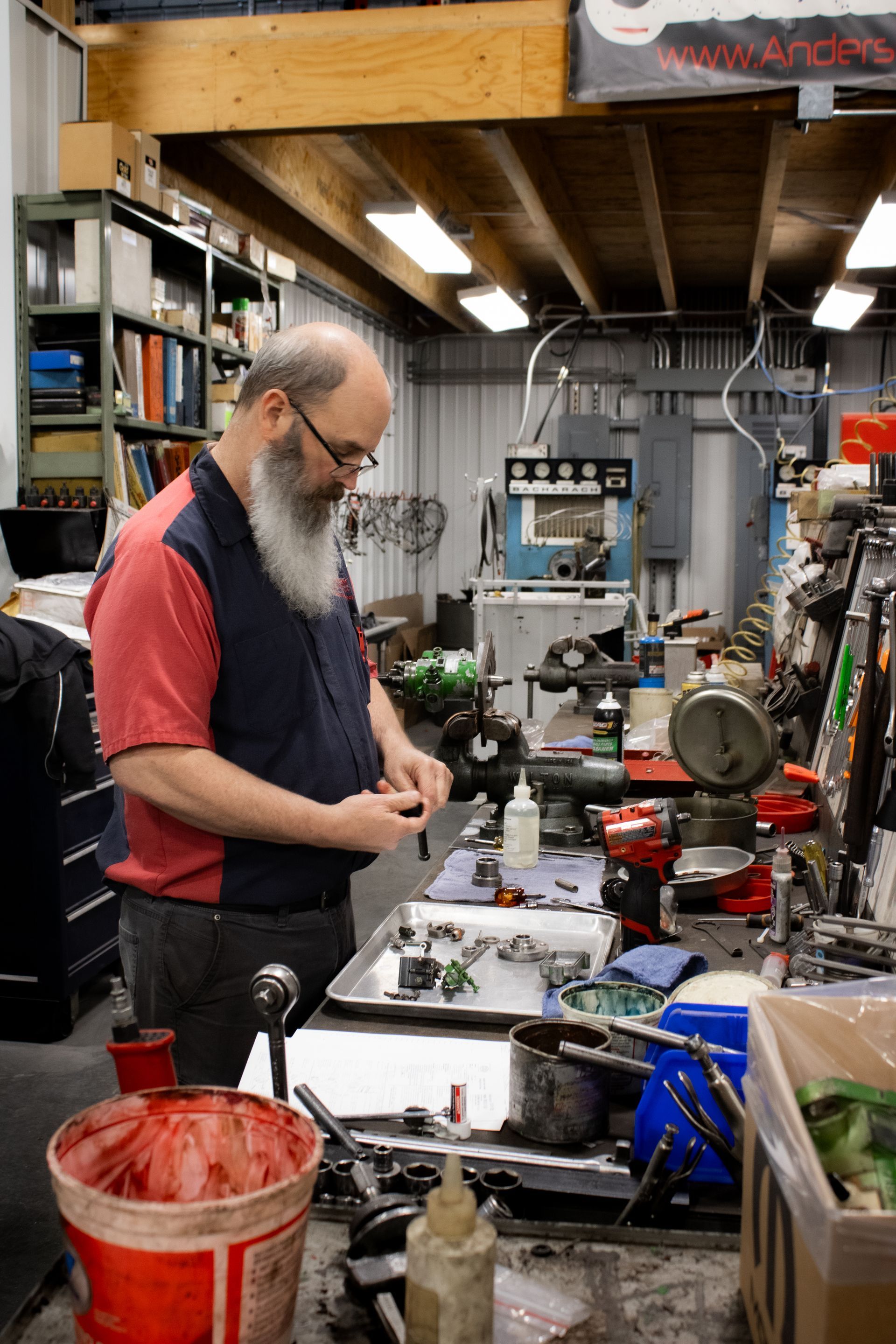 A man working in a cluttered workshop, focused on a task at a bench with tools and equipment.