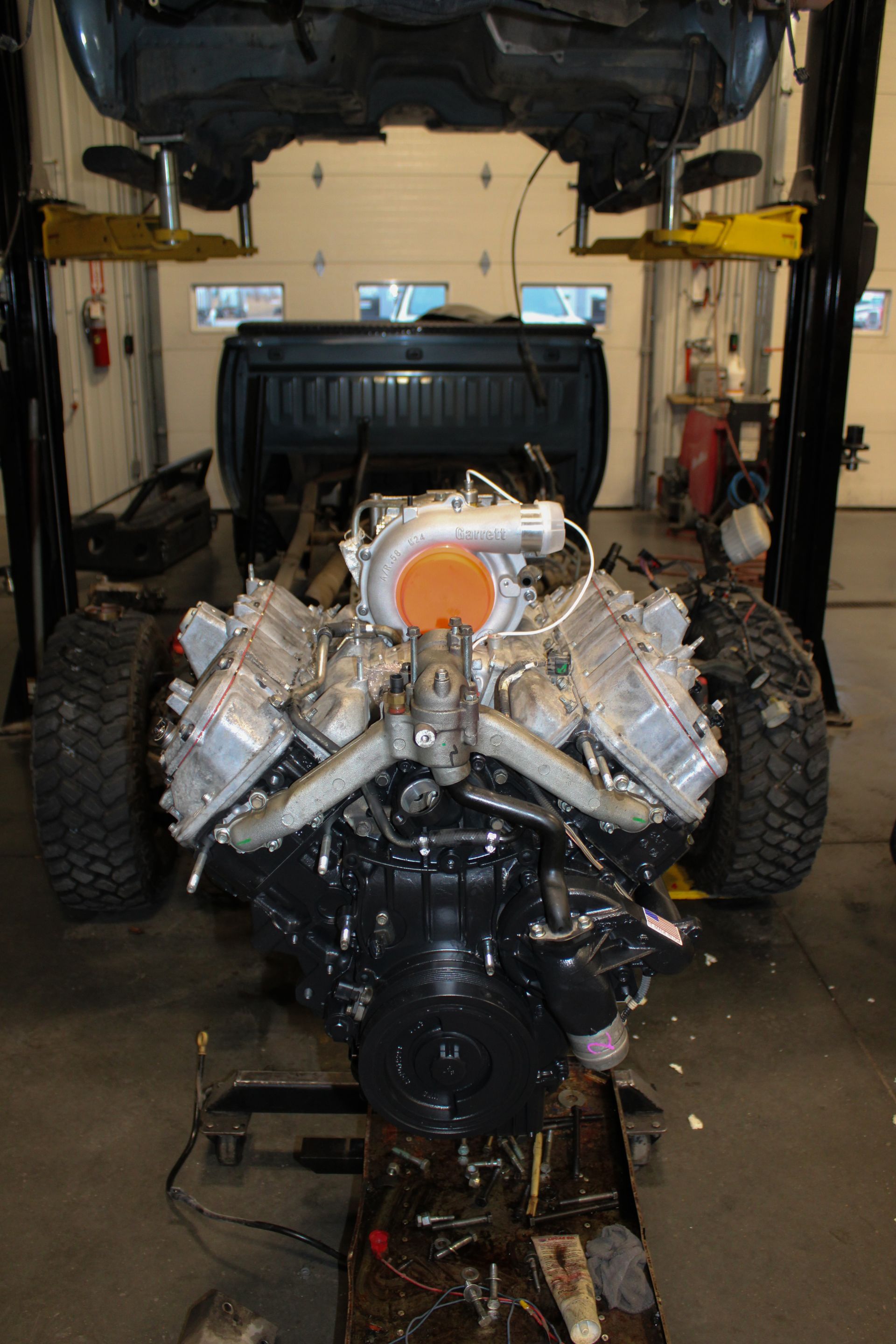Car engine on a stand in a garage, with the hood of a vehicle raised above it.
