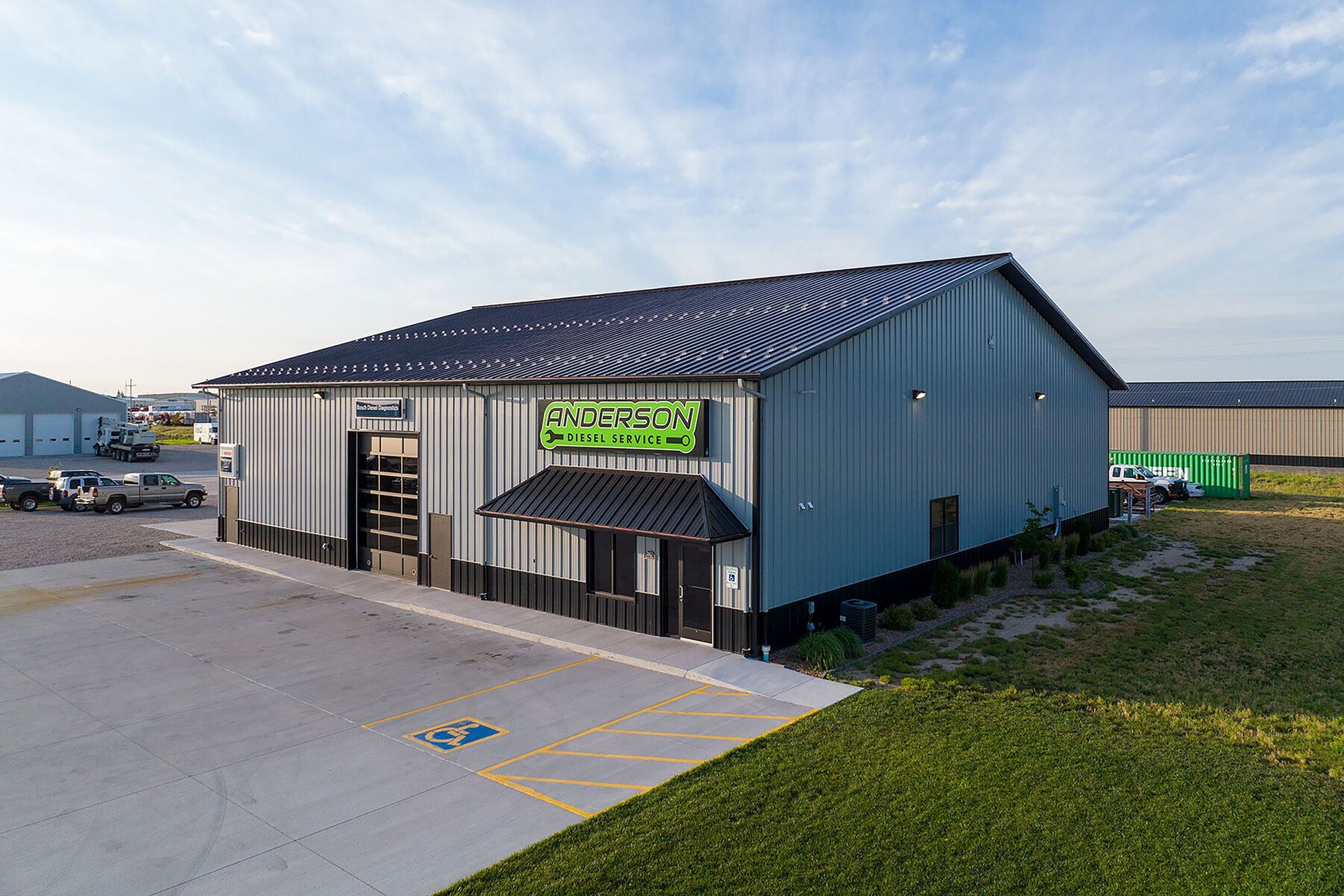An aerial view of a gray, single-story industrial building with a green business sign and a paved parking lot.