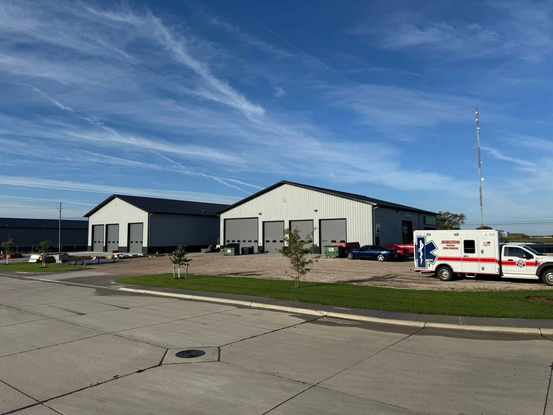 Two white metal industrial buildings under a blue sky, with an ambulance parked in front on a gravel lot.