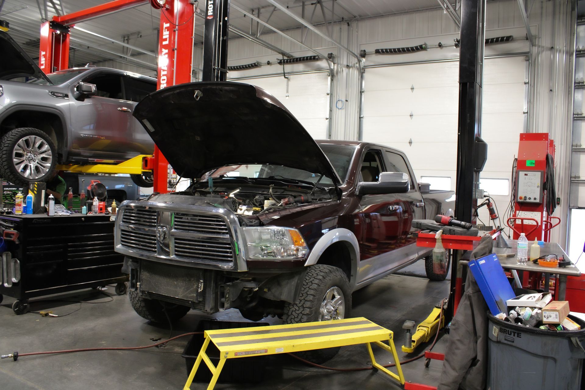 A dark-colored pickup truck with its hood open in an auto repair shop with another car lifted in the background.