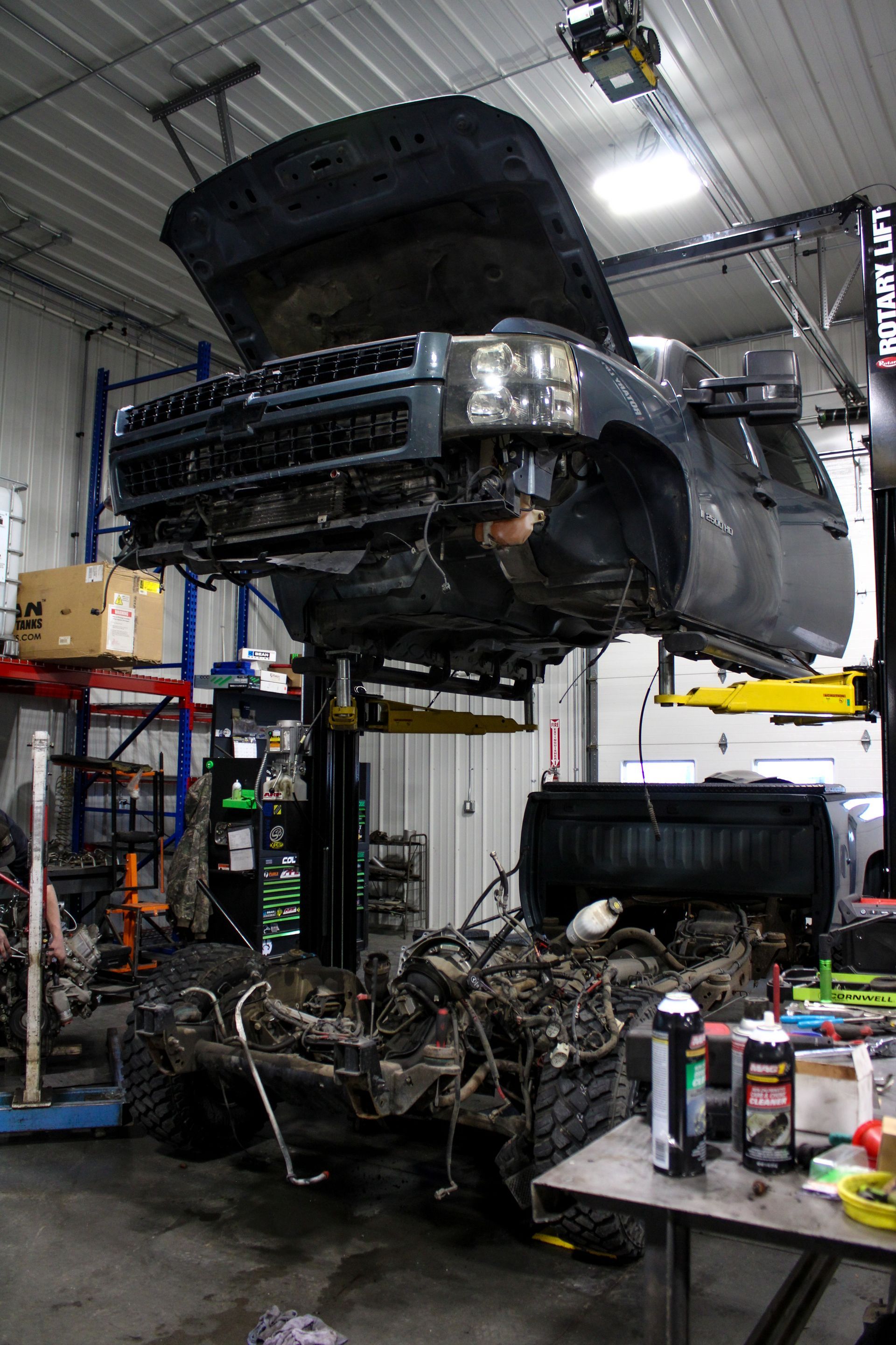 A vehicle body is lifted on a mechanical hoist in a repair shop, exposing the chassis and components on the floor below.