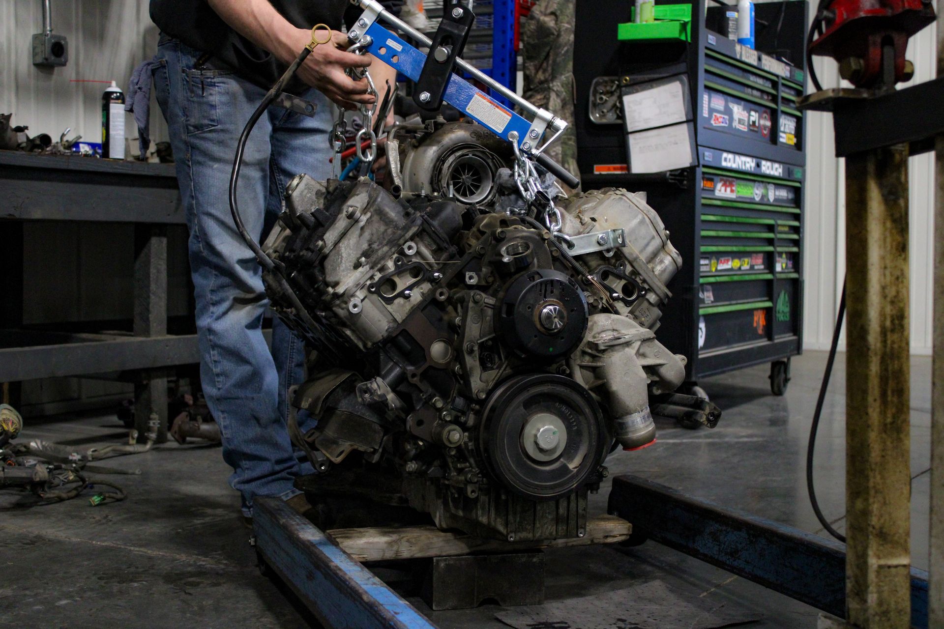 A mechanic guides an engine suspended by a blue lifting bar in a workshop.