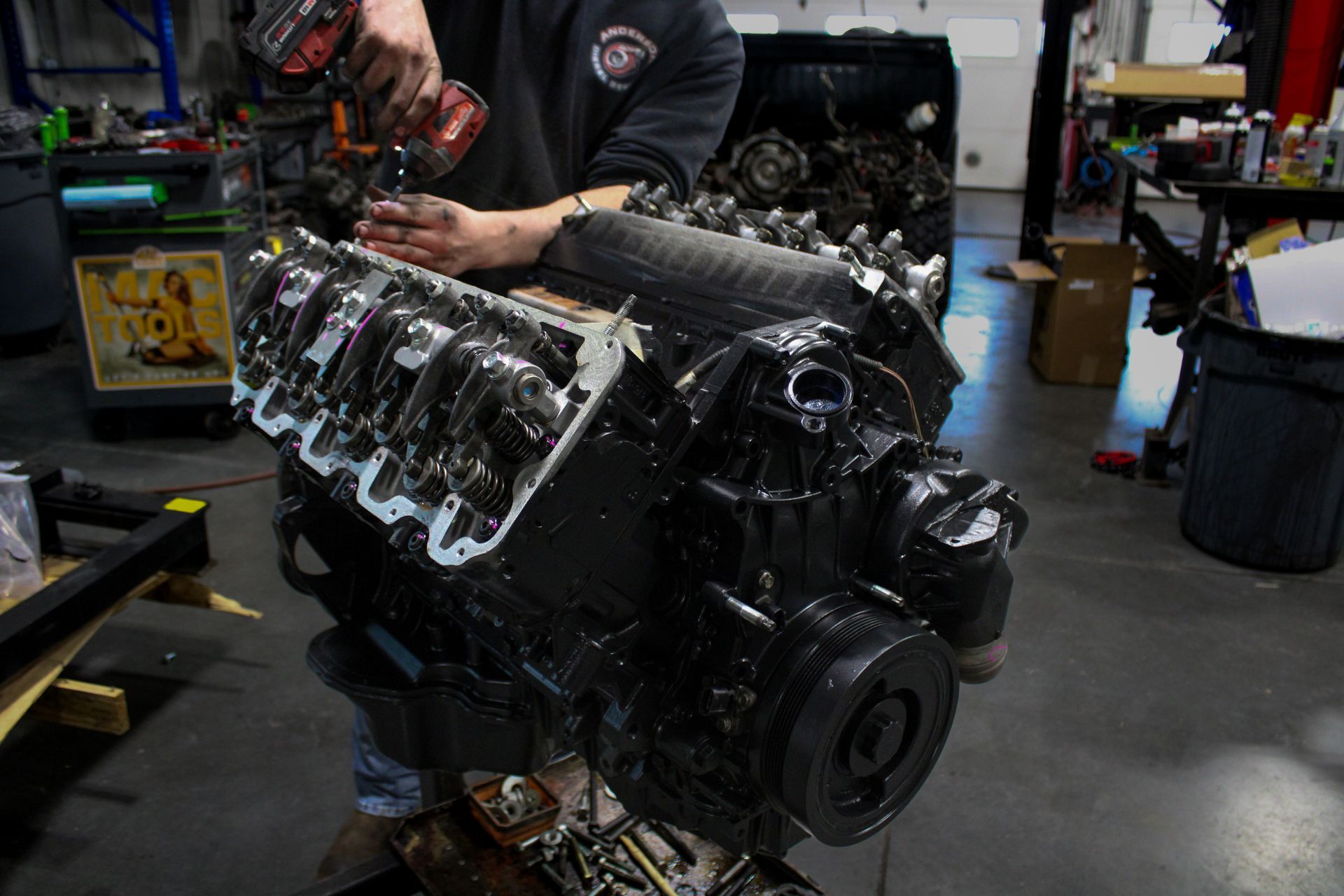 A person uses a power drill on the cylinder head of a black engine block in a busy automotive repair shop.