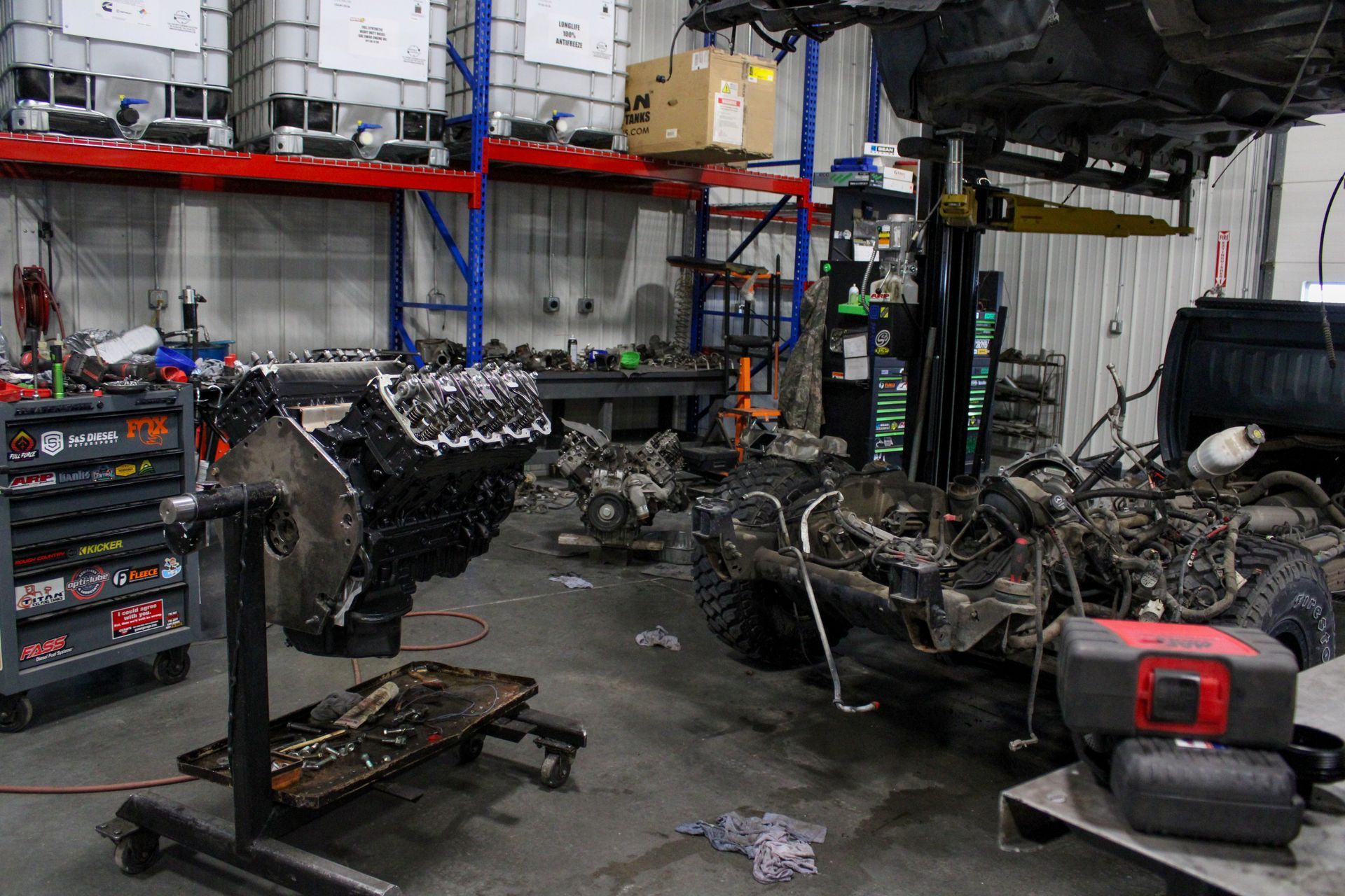 An engine on a stand and a vehicle chassis in a cluttered industrial workshop with shelving units and tools.