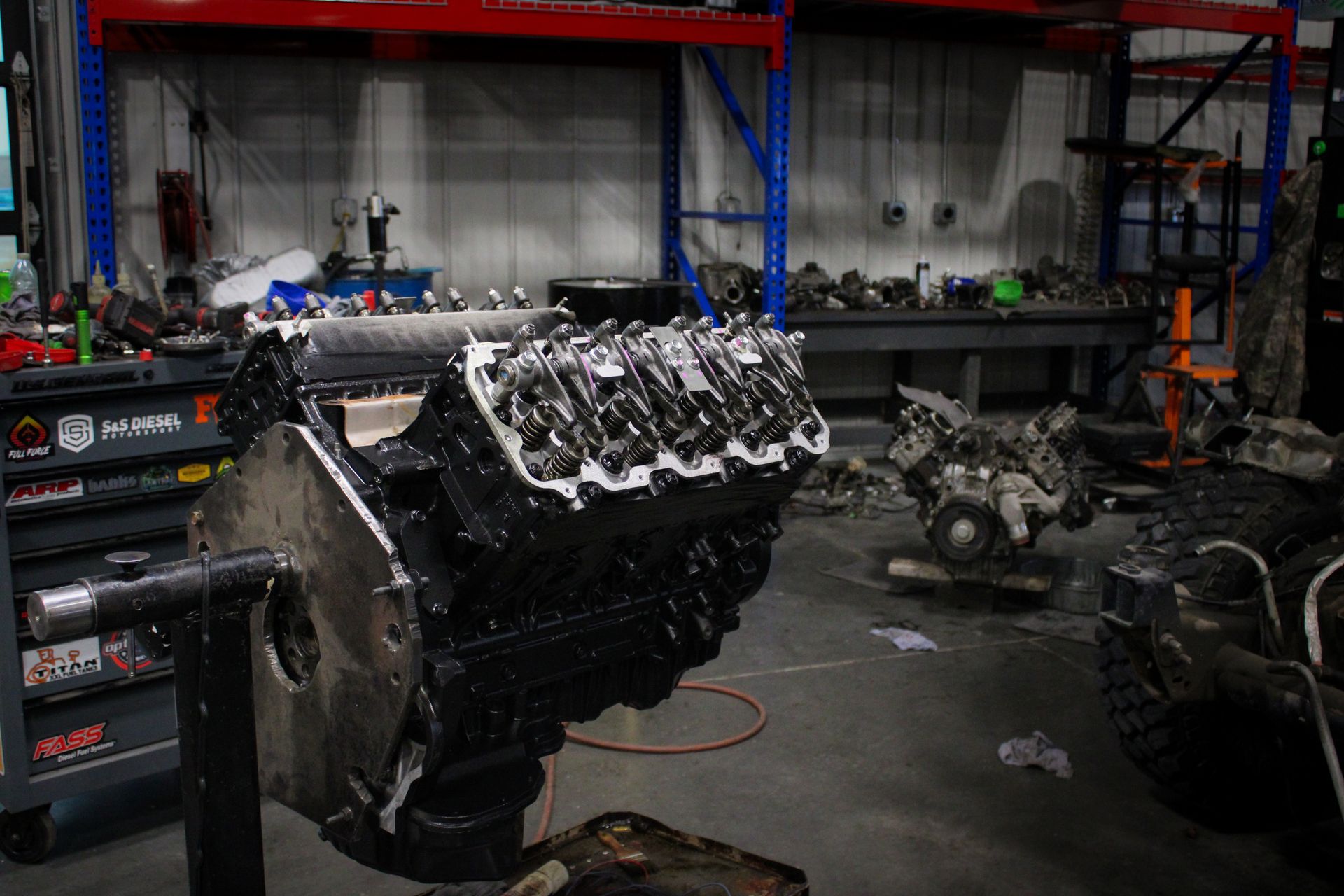 A partially assembled black V8 engine mounted on a stand in a workshop with shelves, tools, and automotive parts.