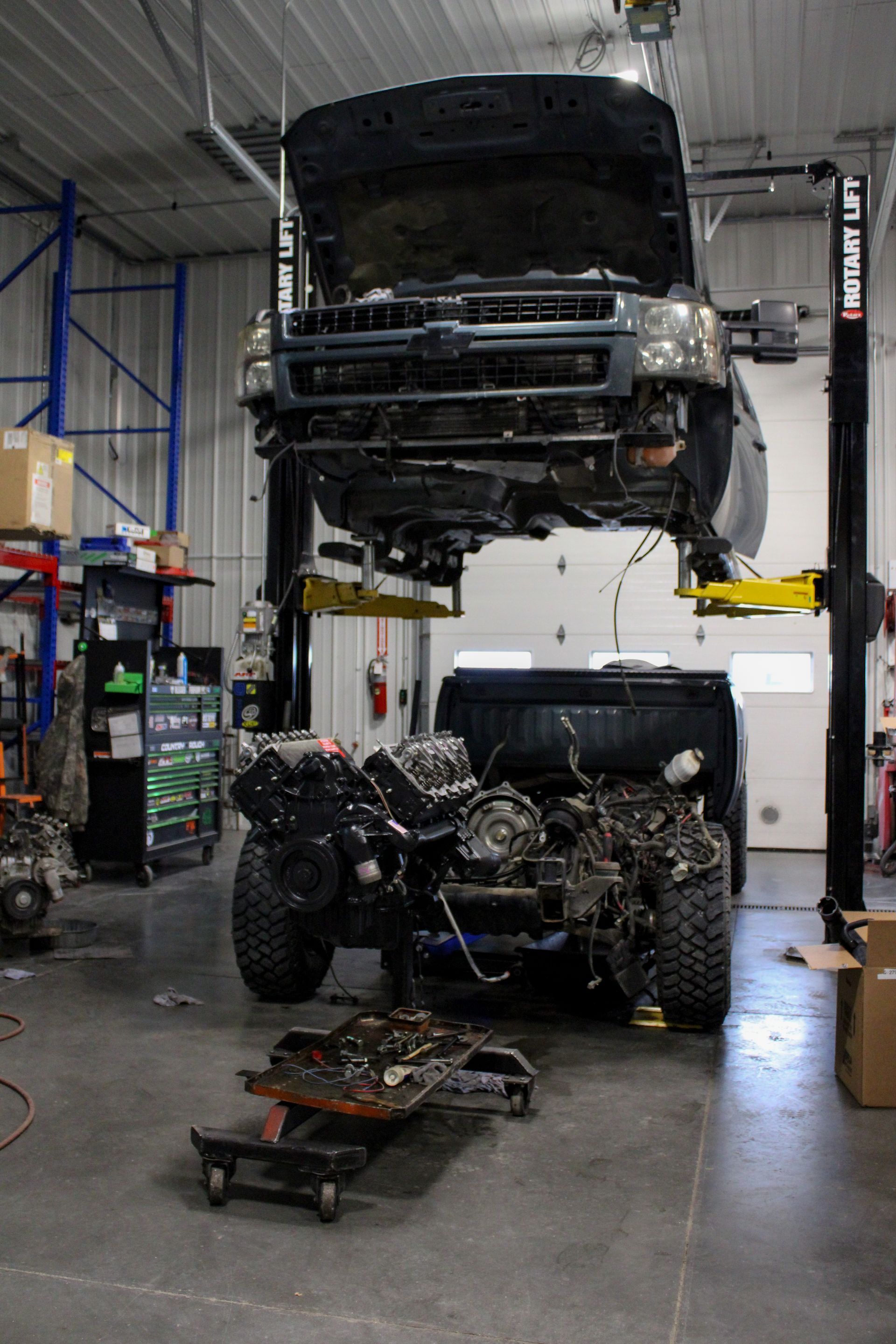 A dark-colored pickup truck is raised on a hydraulic auto lift in a garage, with its engine removed and resting below.